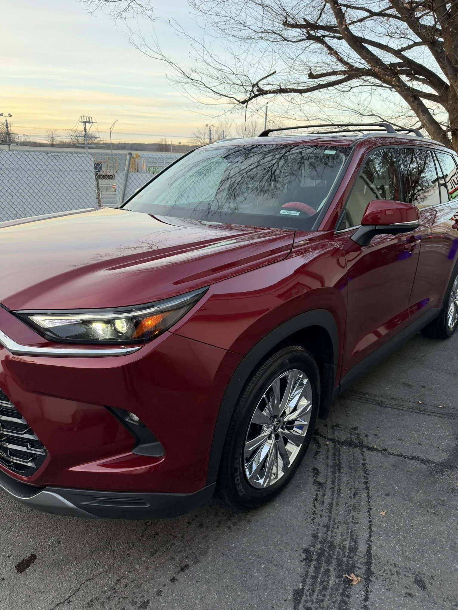A red SUV parked on a paved lot with a chain-link fence, leafless trees, and a sunset sky in the background.