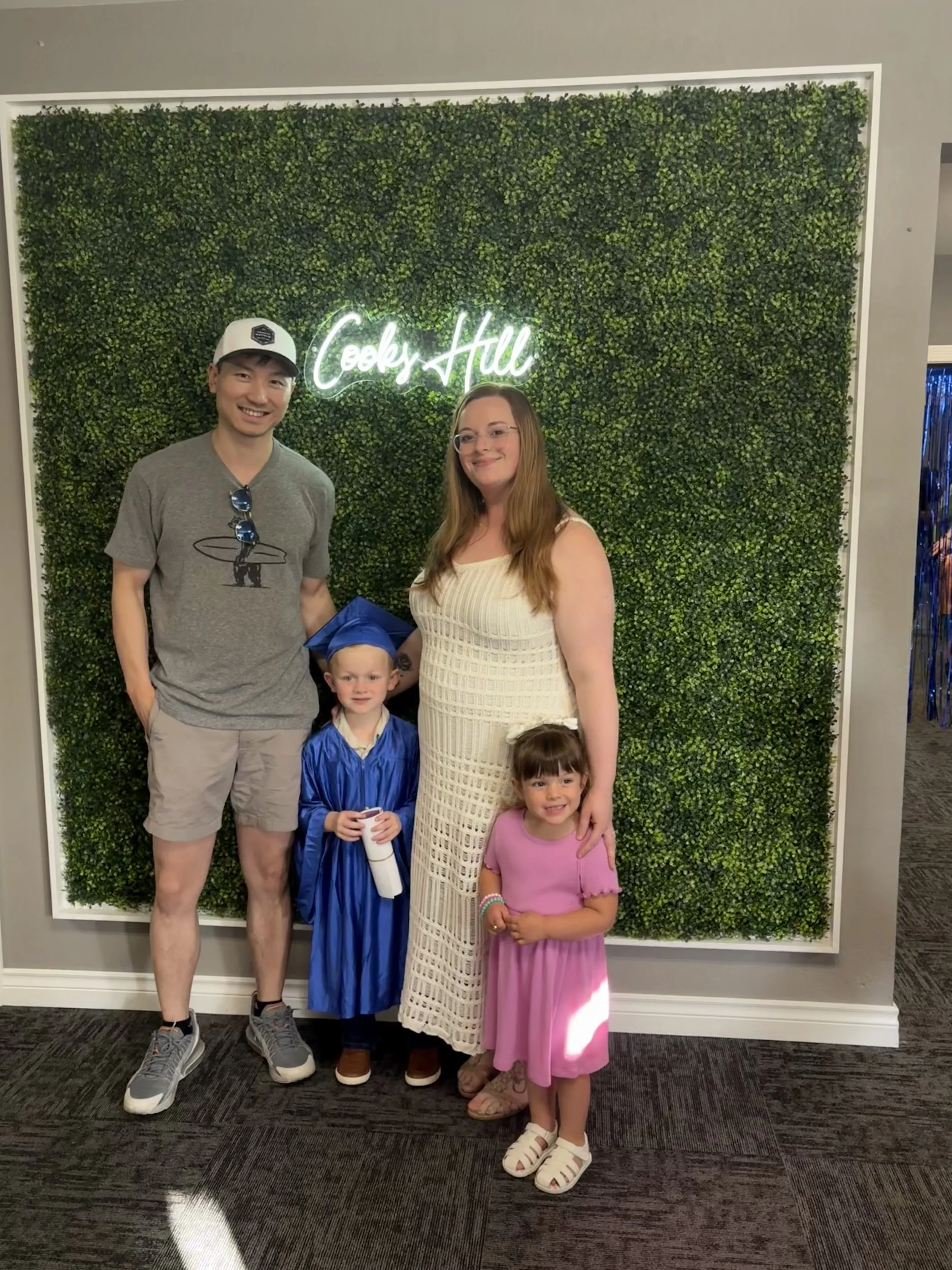 Family celebrating a graduation at Cook's Hill, standing in front of a green wall with a neon sign that says "Cook's Hill." A young boy in a blue graduation gown and cap holds a diploma, flanked by a woman in a cream dress and two young girls in pink dresses.
