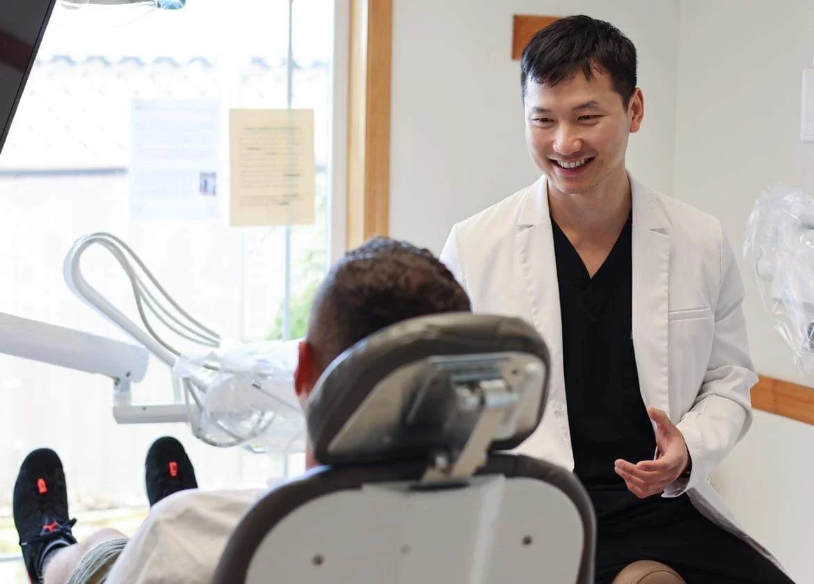 A young male dentist dressed in a white coat smiling and talking to a patient in a dental chair.