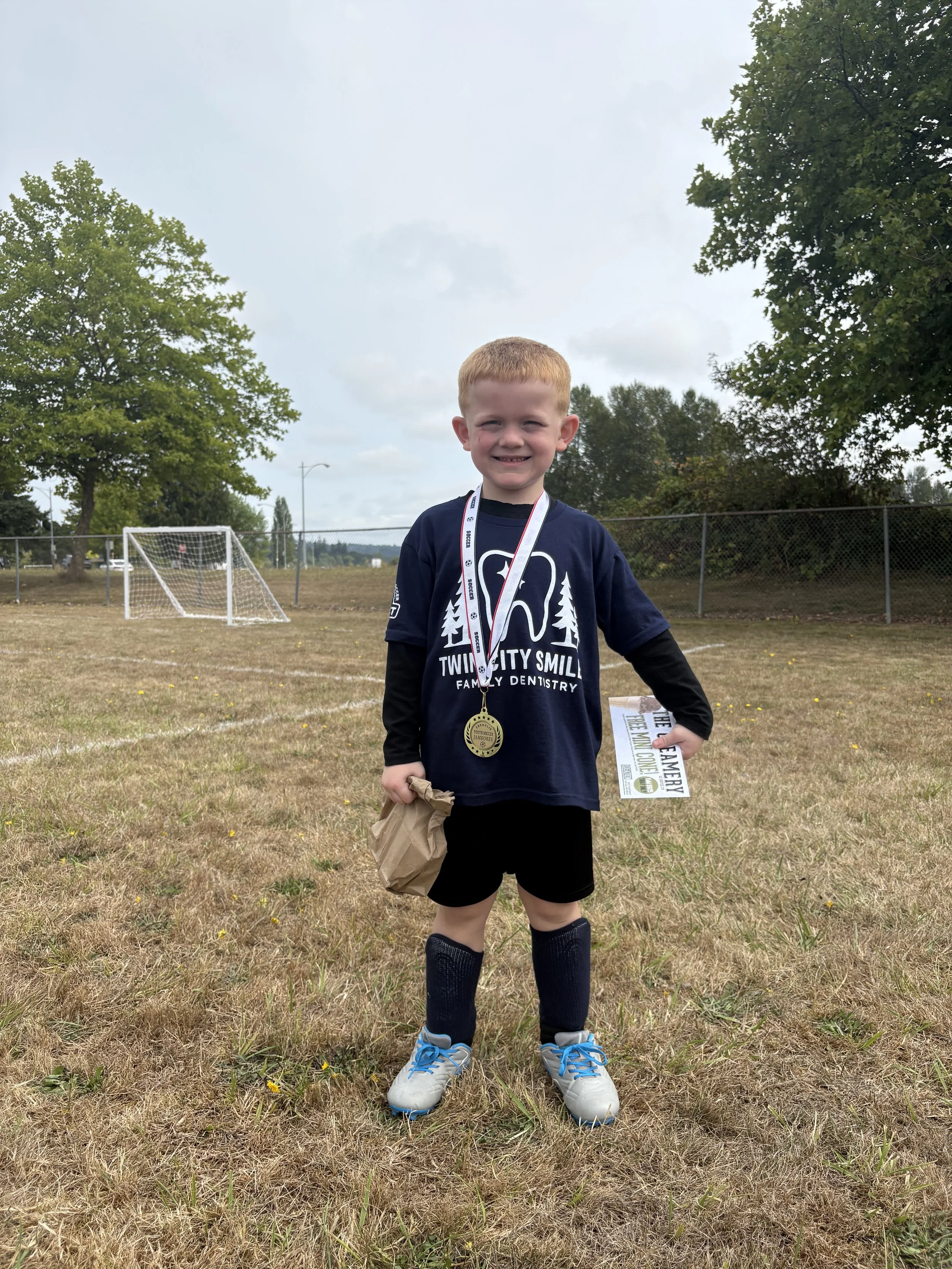 Young boy wearing a sports uniform with medals around his neck, standing on a field with a soccer goal in the background, holding a certificate and a paper bag.
