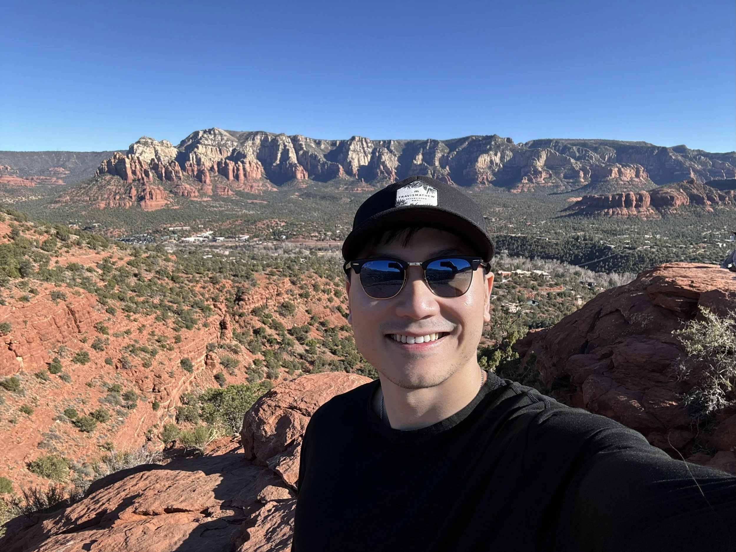 A smiling man wearing sunglasses and a black cap taking a selfie in a desert landscape with red rocks and mountains in the background