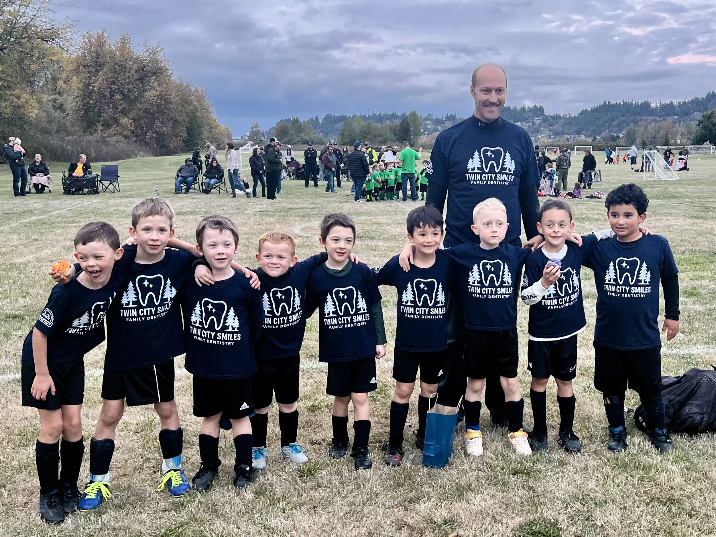 A group of young boys in soccer uniforms with a coach in a field after a game, with other people and children in the background.