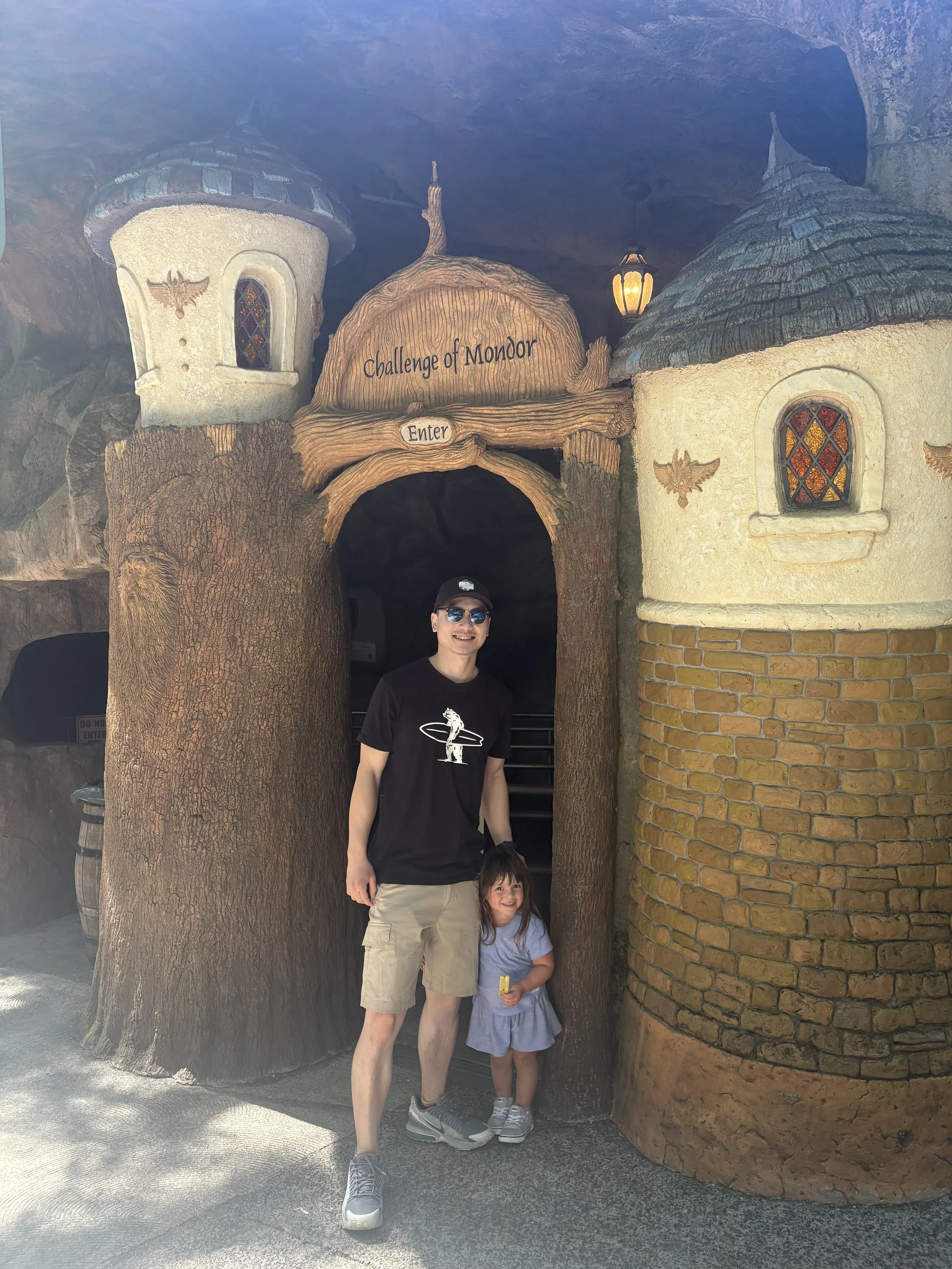 Man and girl standing in front of the entrance to the 'Challenge of Morder' ride, with fairy tale castle-like structures in the background.