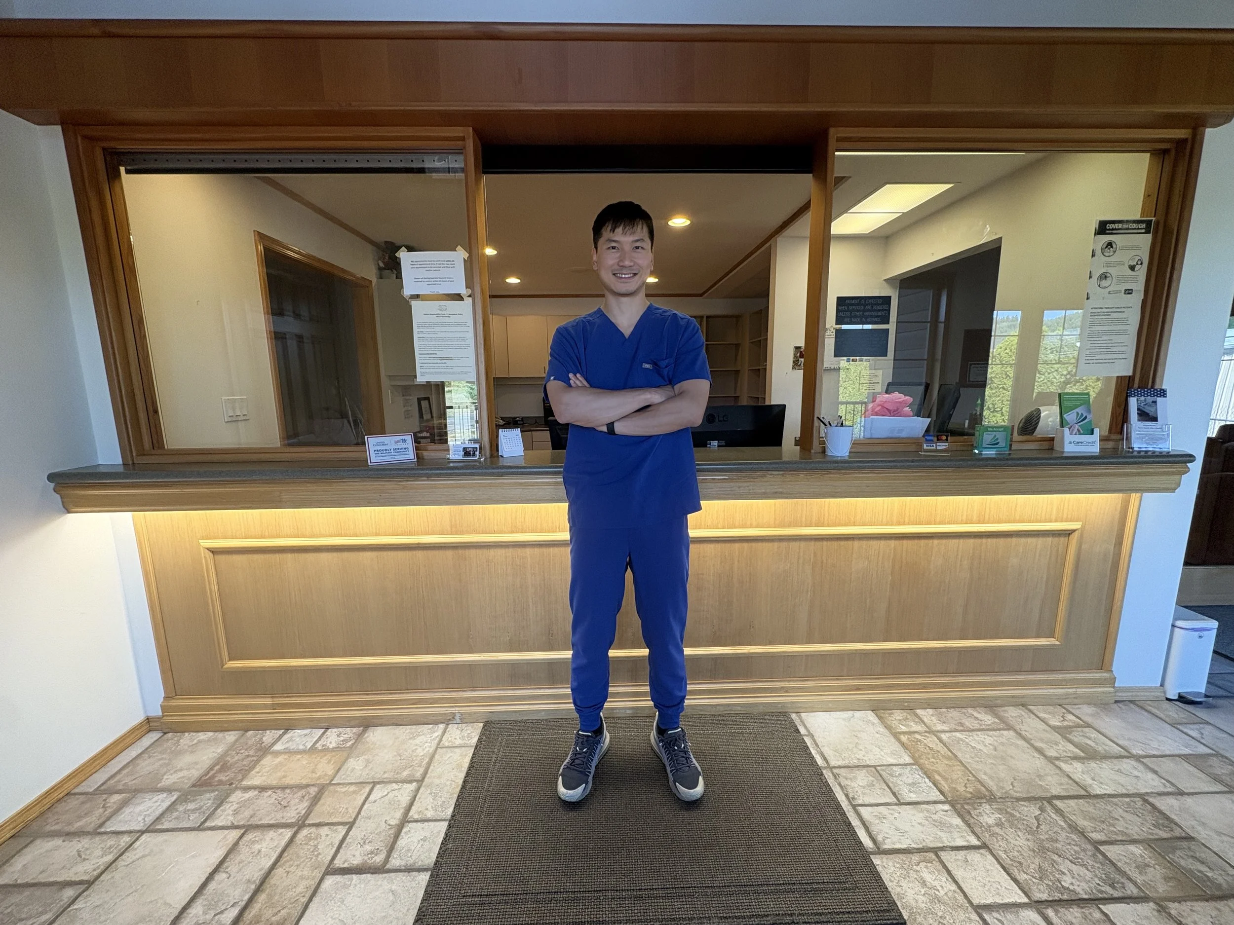Male dentist in blue scrubs standing with arms crossed at Twin City Smiles reception desk in Centralia, WA