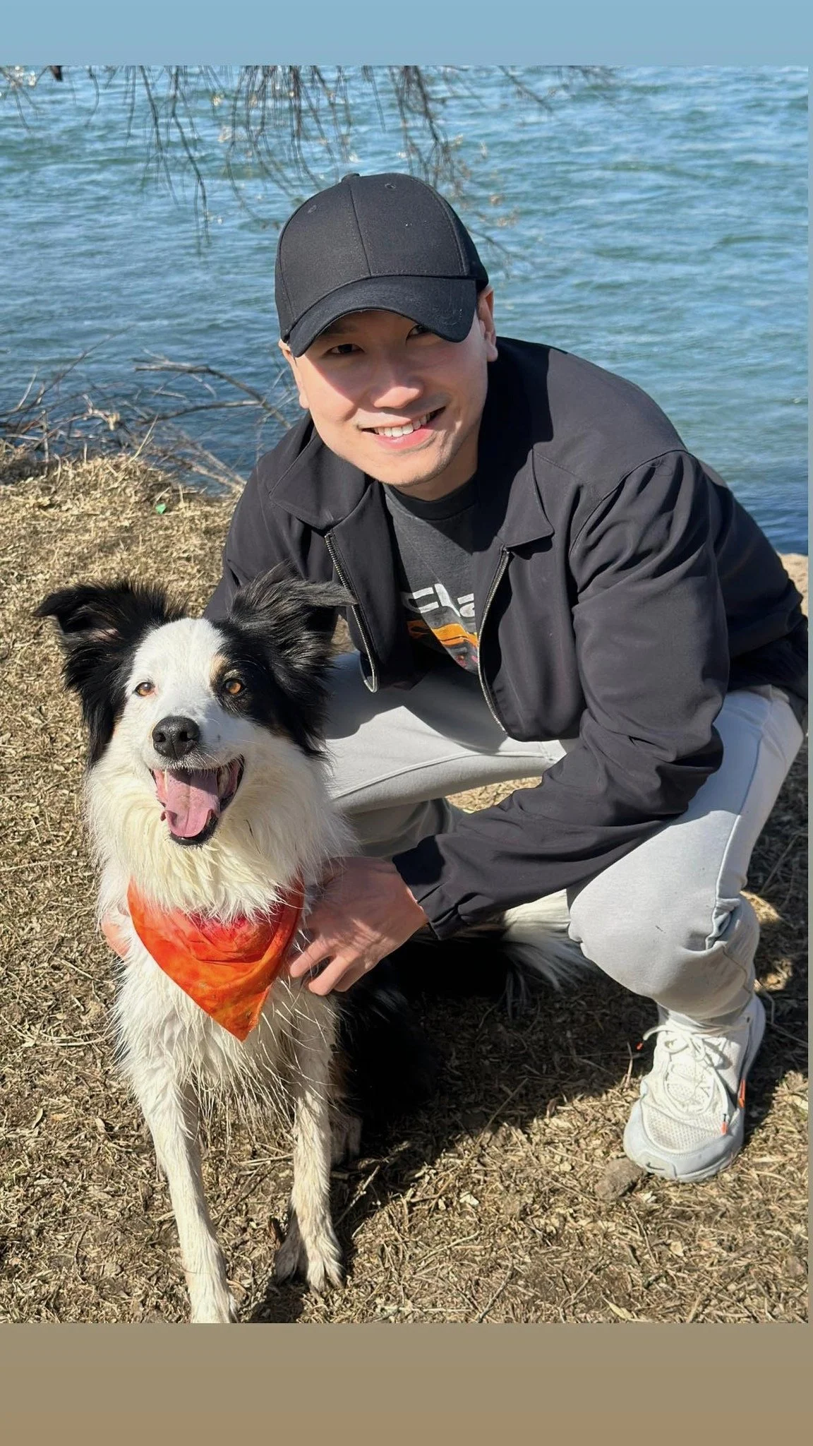 A man wearing a black cap and black jacket crouching down beside a happy black and white border collie dog with an orange bandana, near a body of water on a sunny day.