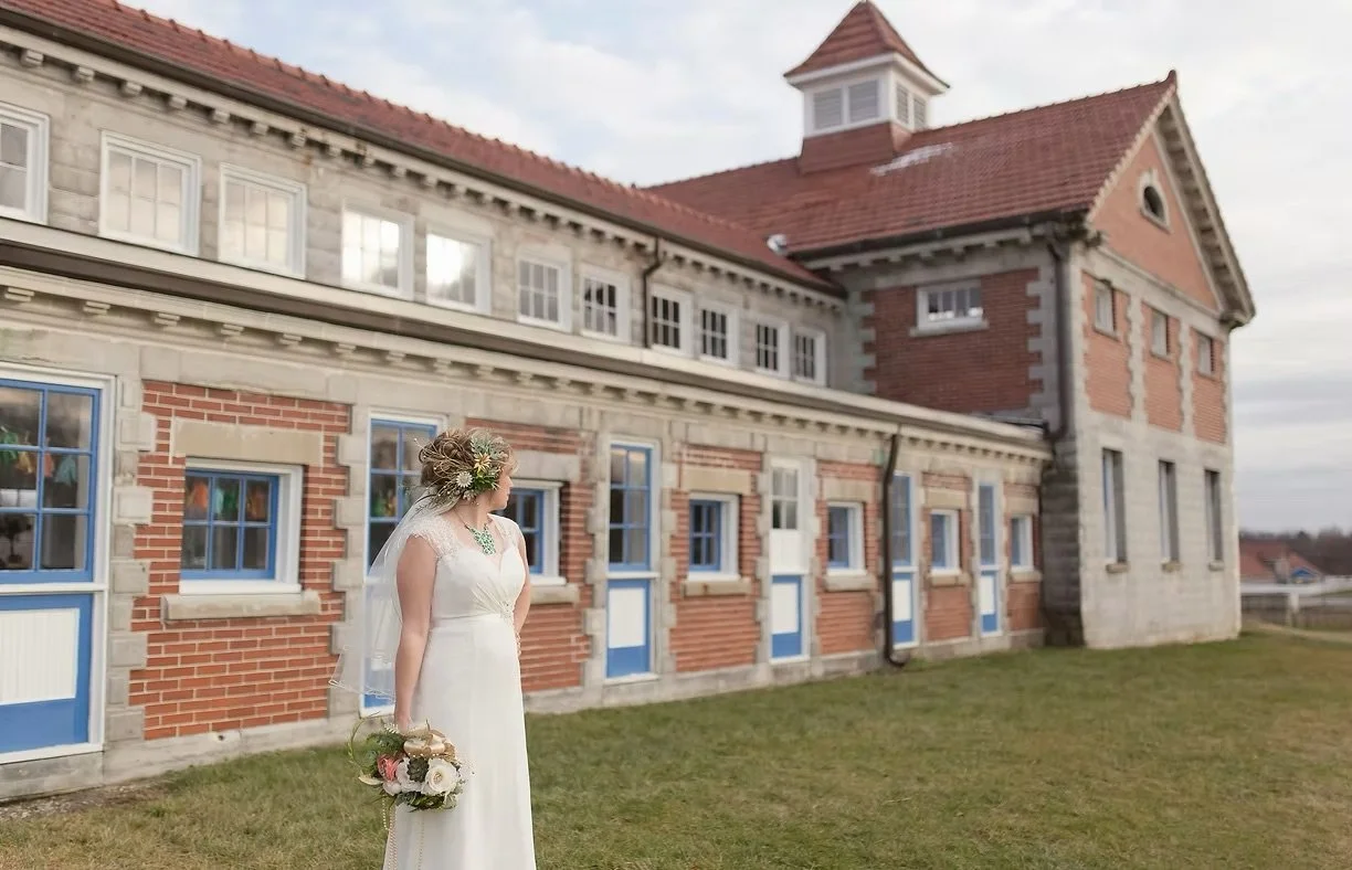 A bride stands on grass outside a large brick and stone building with blue window frames, holding a bouquet of flowers, during daytime.