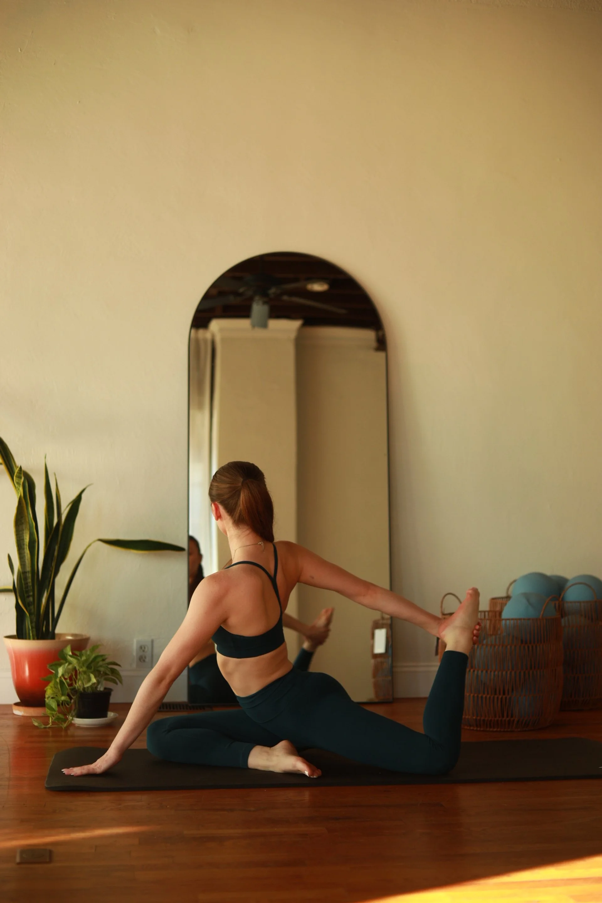 A woman practicing yoga, sitting on a black mat on wooden floor, reaching back to hold her foot in a seated twist pose, in a room with a large mirror, potted plants, and a basket of yoga blocks.