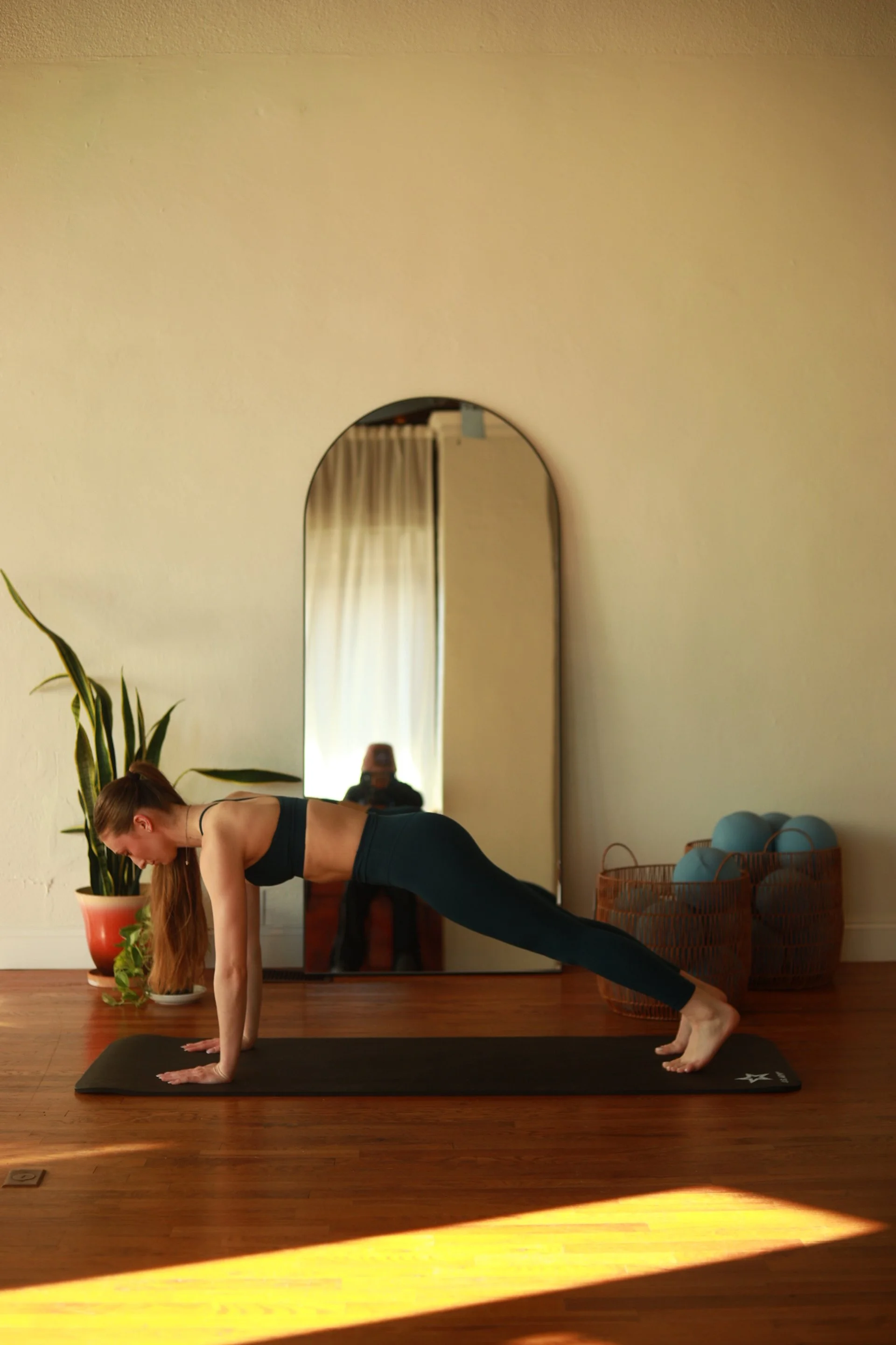 A woman practicing yoga, seated on a mat, with legs extended and arms reaching forward in a forward bend pose, in a room with wooden flooring.