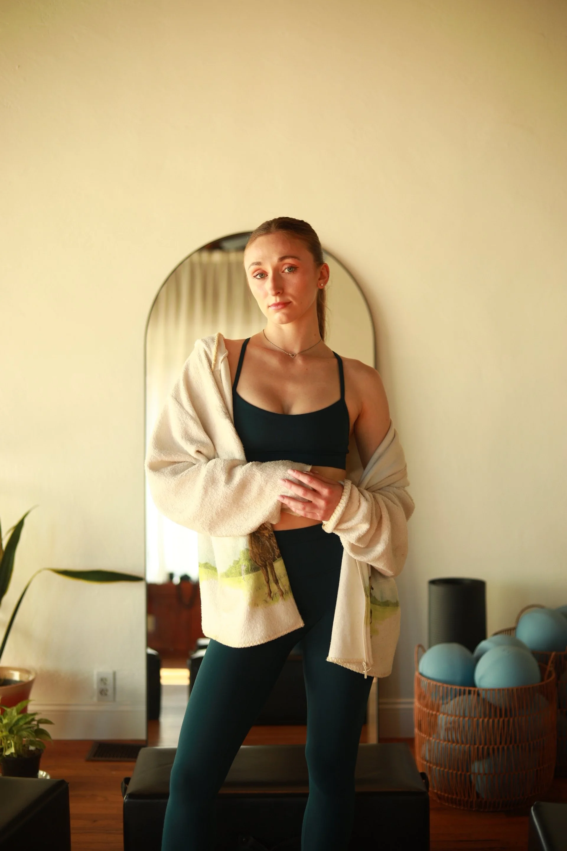 A young woman standing in front of a mirror in a room with wooden flooring and a basket of blue balls. She is wearing a black sports bra, black leggings, and a beige jacket draped over her shoulders, with her hair tied back.