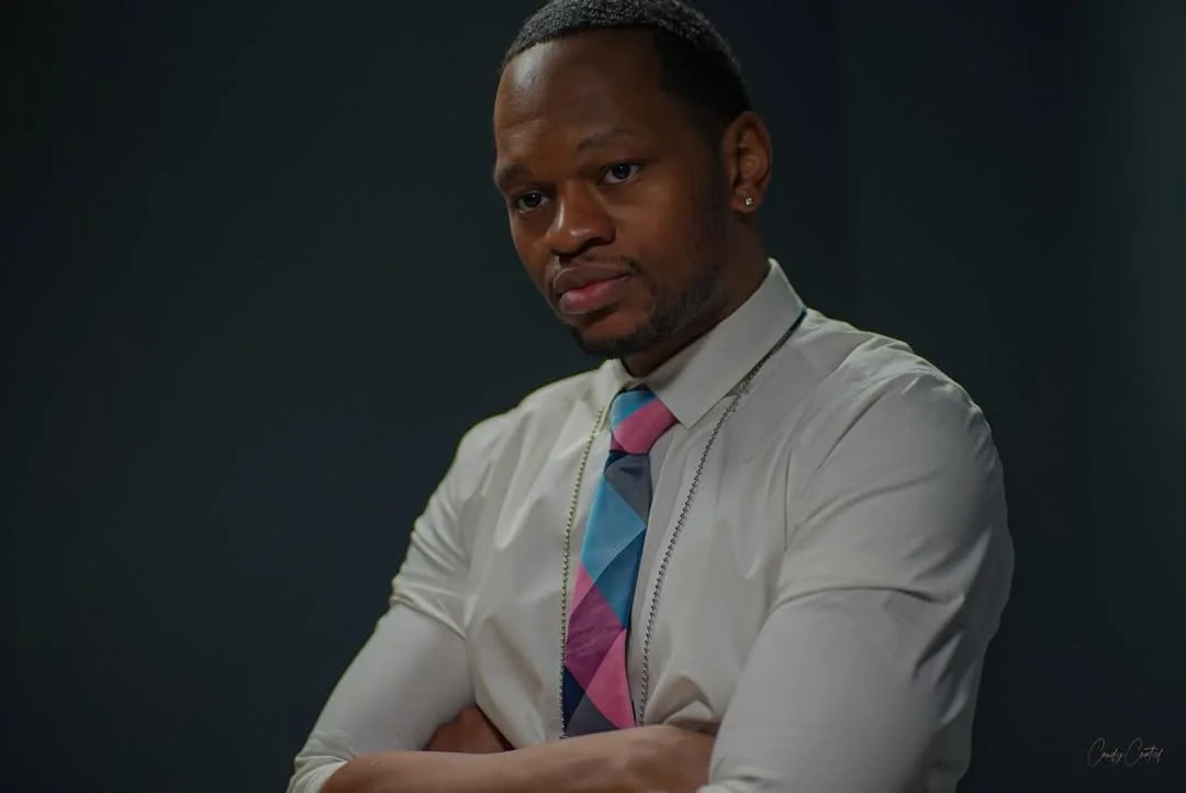A man with crossed arms wearing a white shirt, colorful tie, and earrings, looking at the camera against a dark background.