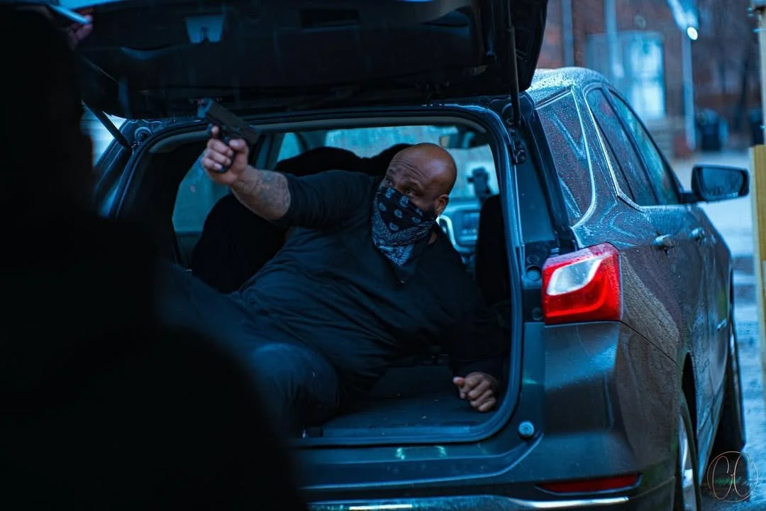 A man wearing a black shirt and bandana mask is inside the trunk of a gray SUV, pointing a handgun at an unseen person.