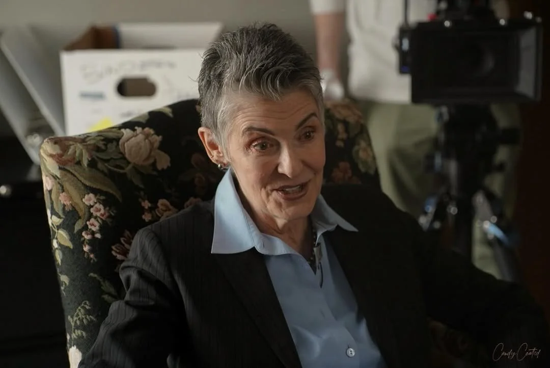 An older woman with short gray hair, wearing a black blazer and a light blue blouse, sitting in a floral armchair, speaking or laughing.