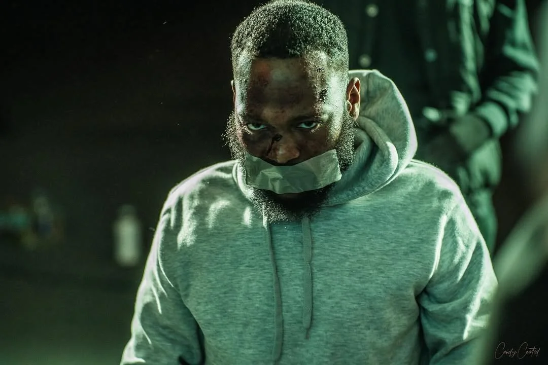 A young man with a beard and short hair, wearing a gray hoodie and a face mask, looking intently at the camera, with a dark background and another person partially visible in the background.