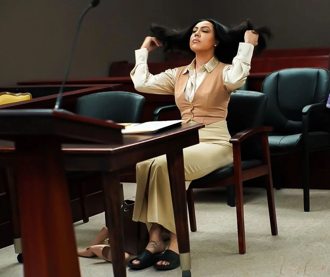 A woman in professional attire sitting in a courtroom or conference room, with her eyes closed and holding her hair back.