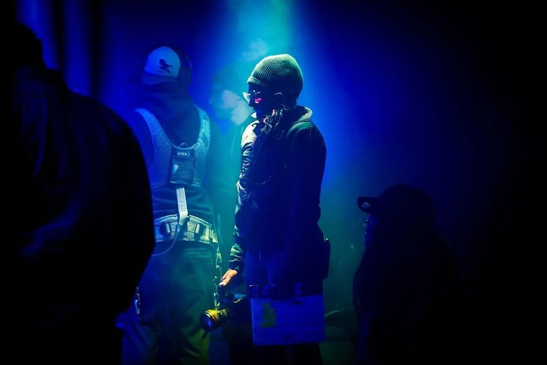 Group of people in dark clothing, some wearing jackets and hats, standing in blue and green lighting, with one person holding a flashlight.