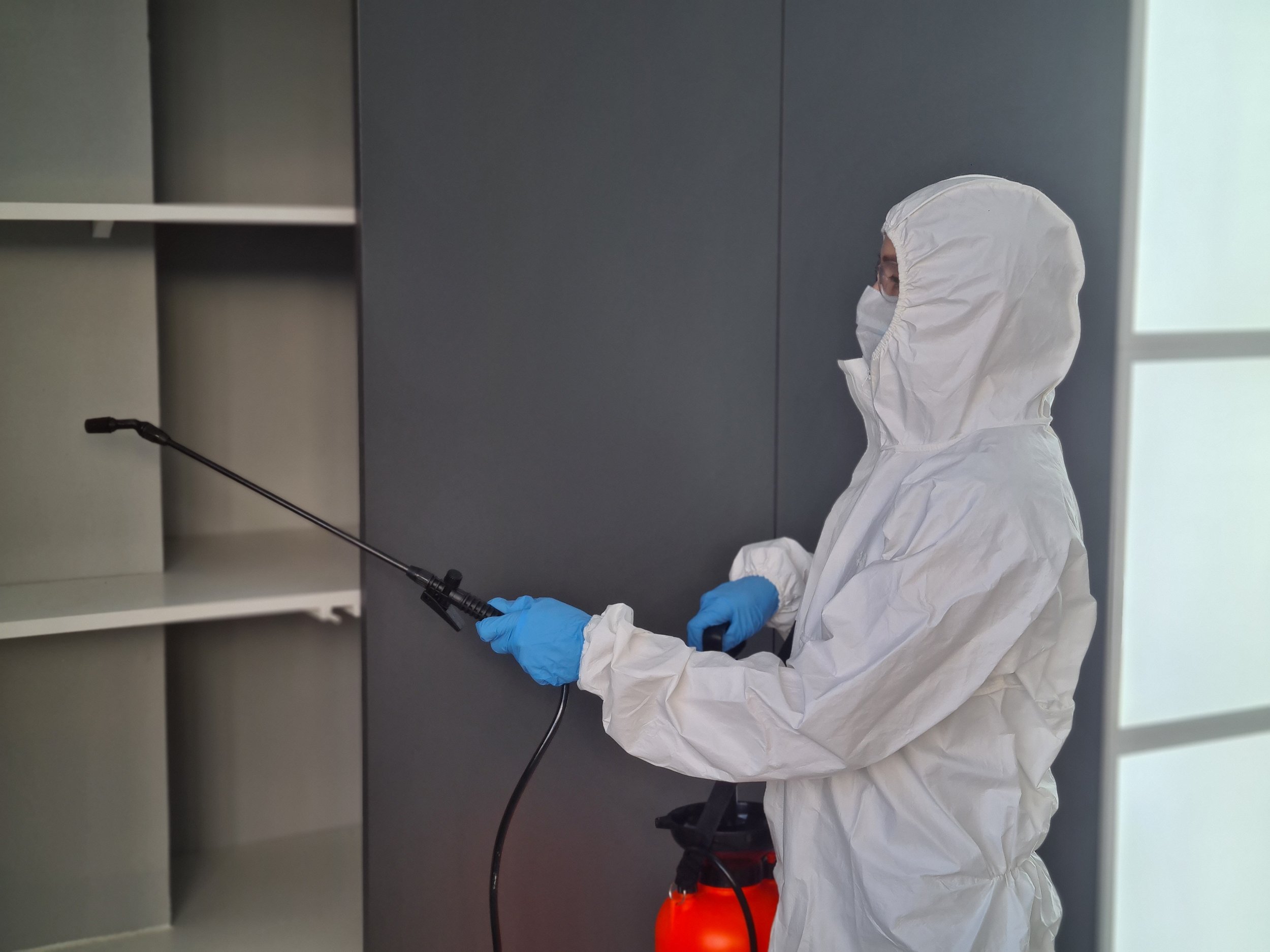 A person in full protective gear, including a white hazmat suit, mask, and blue gloves, is disinfecting or cleaning a surface with a spray nozzle.