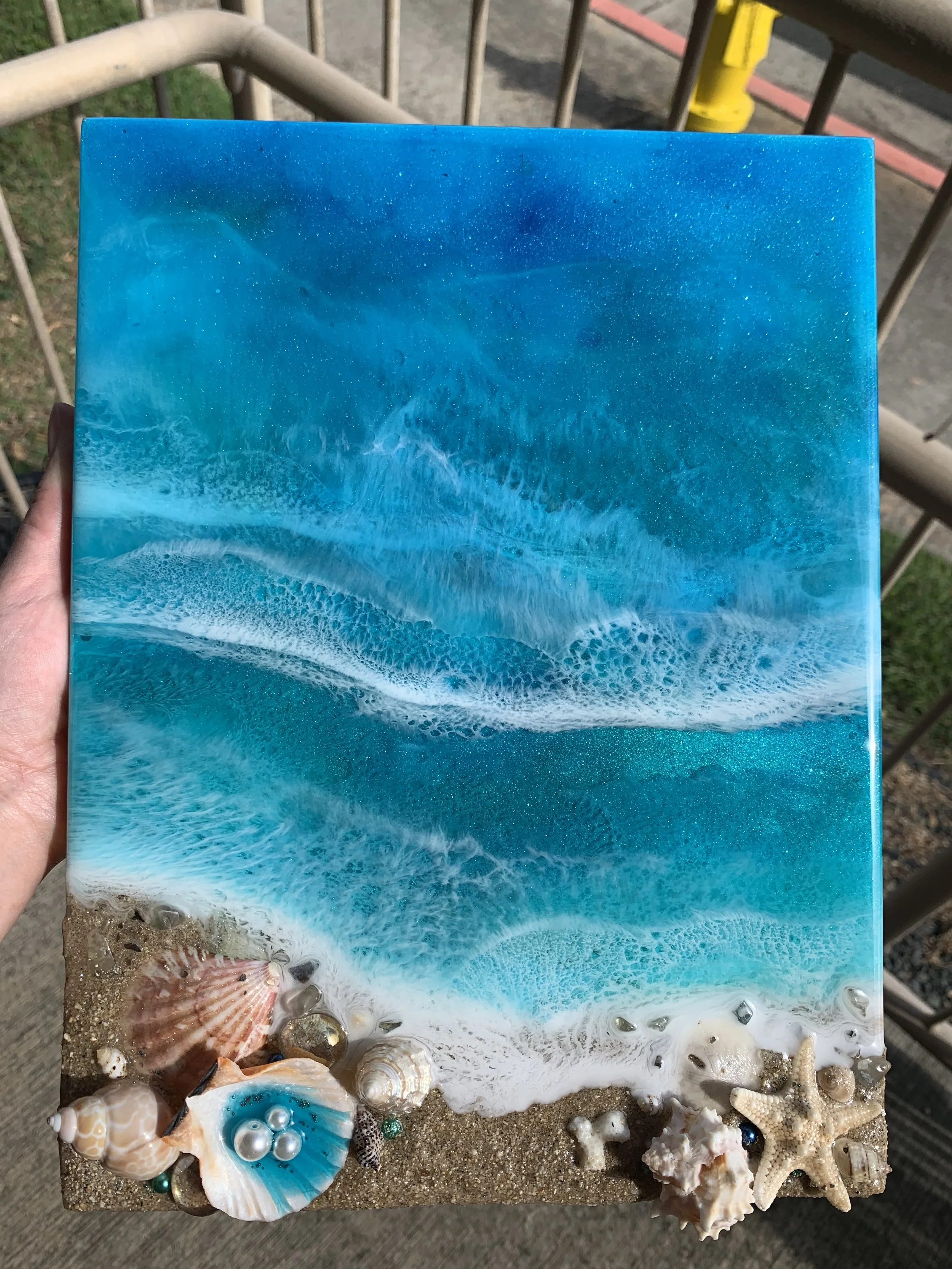 A handcrafted artwork depicts an ocean scene with waves and beach sand, decorated with seashells, starfish, and small decorative stones along the bottom edge.