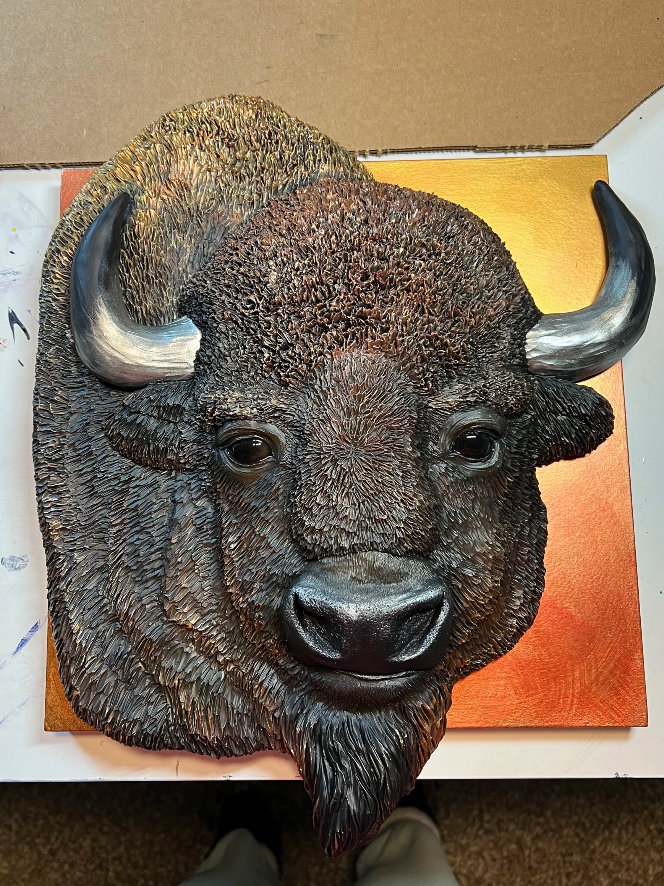 A detailed sculpture of a bison's head with textured fur, prominent black horns, and a shiny black nose, mounted on a golden square background.