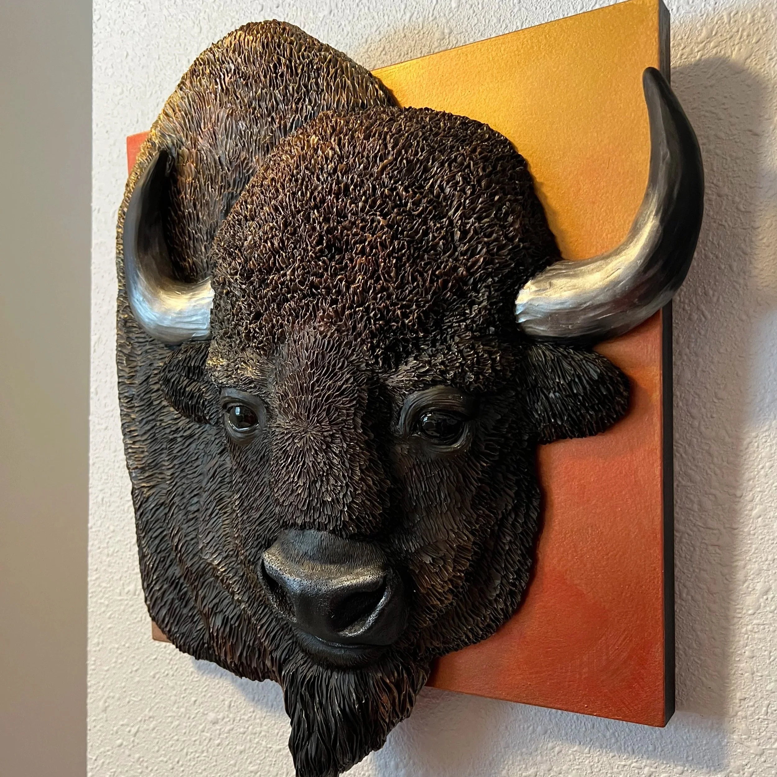 3D wall sculpture of a bison's head with detailed textured fur, large curved horns, and realistic eyes, mounted on a square orange background.