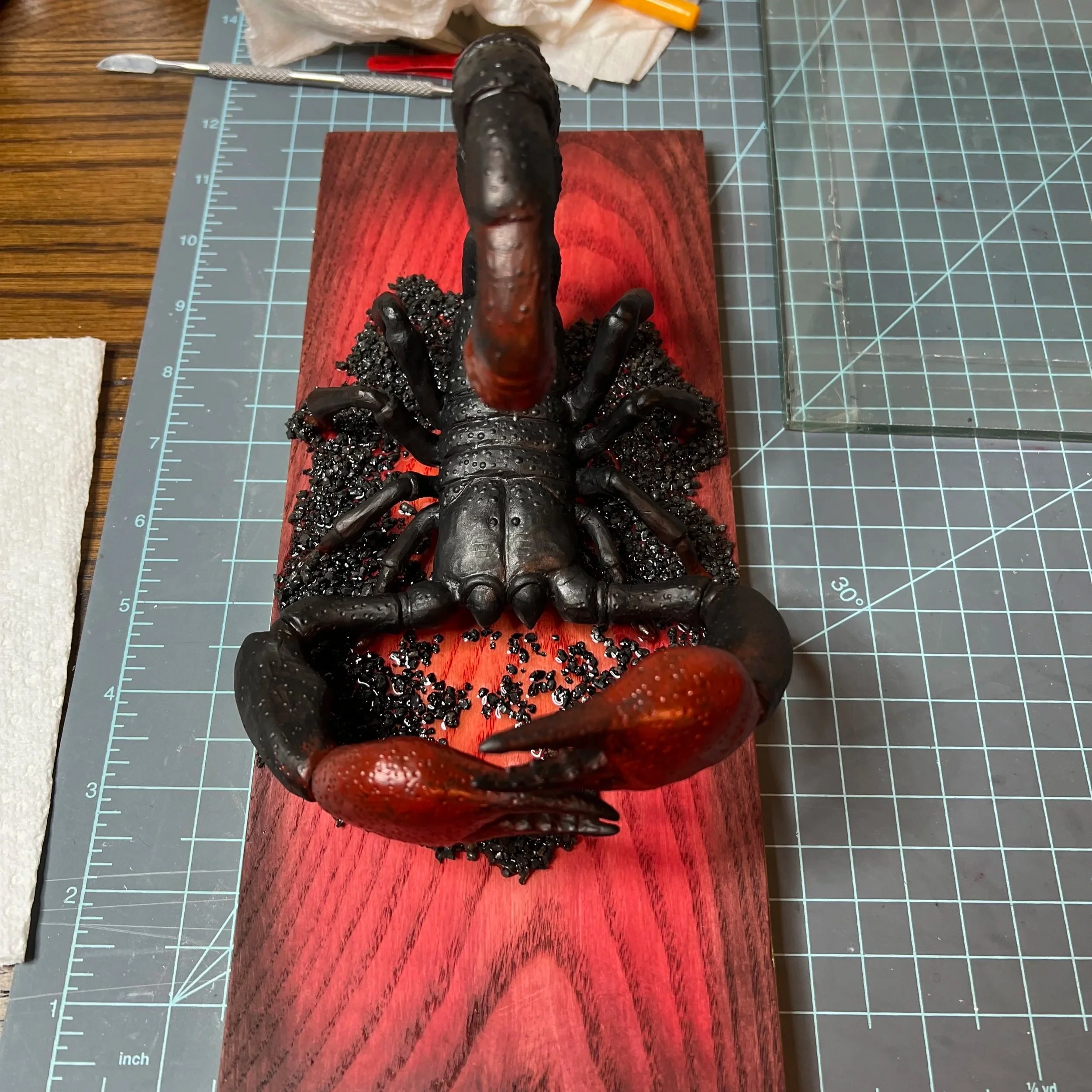 A detailed sculpture of a black and red lobster mounted on a rectangular red wood base, surrounded by small black rocks or gravel, on a crafting mat with tools nearby.