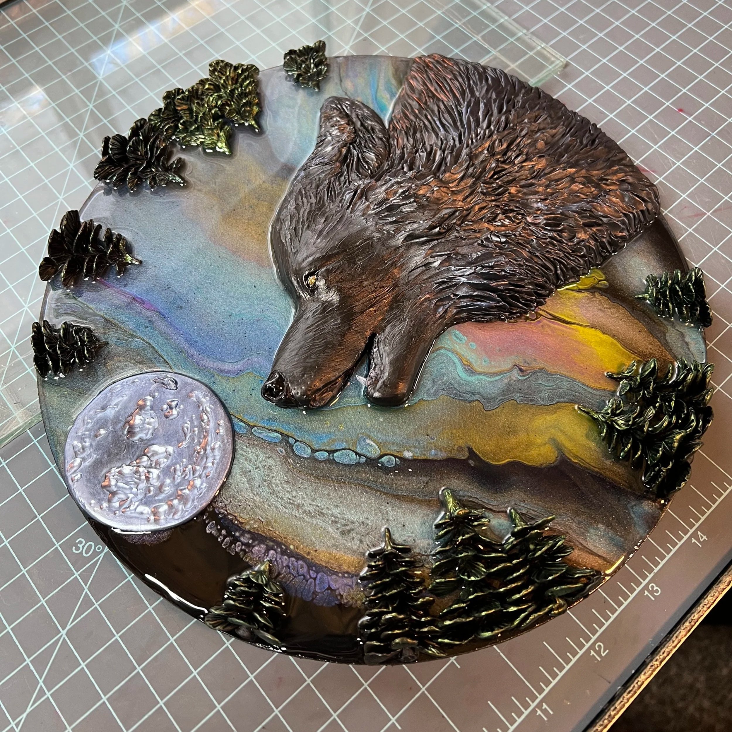 A decorative glass art piece depicting a wolf's head in profile surrounded by pine trees and a moon, with colorful swirling backgrounds.