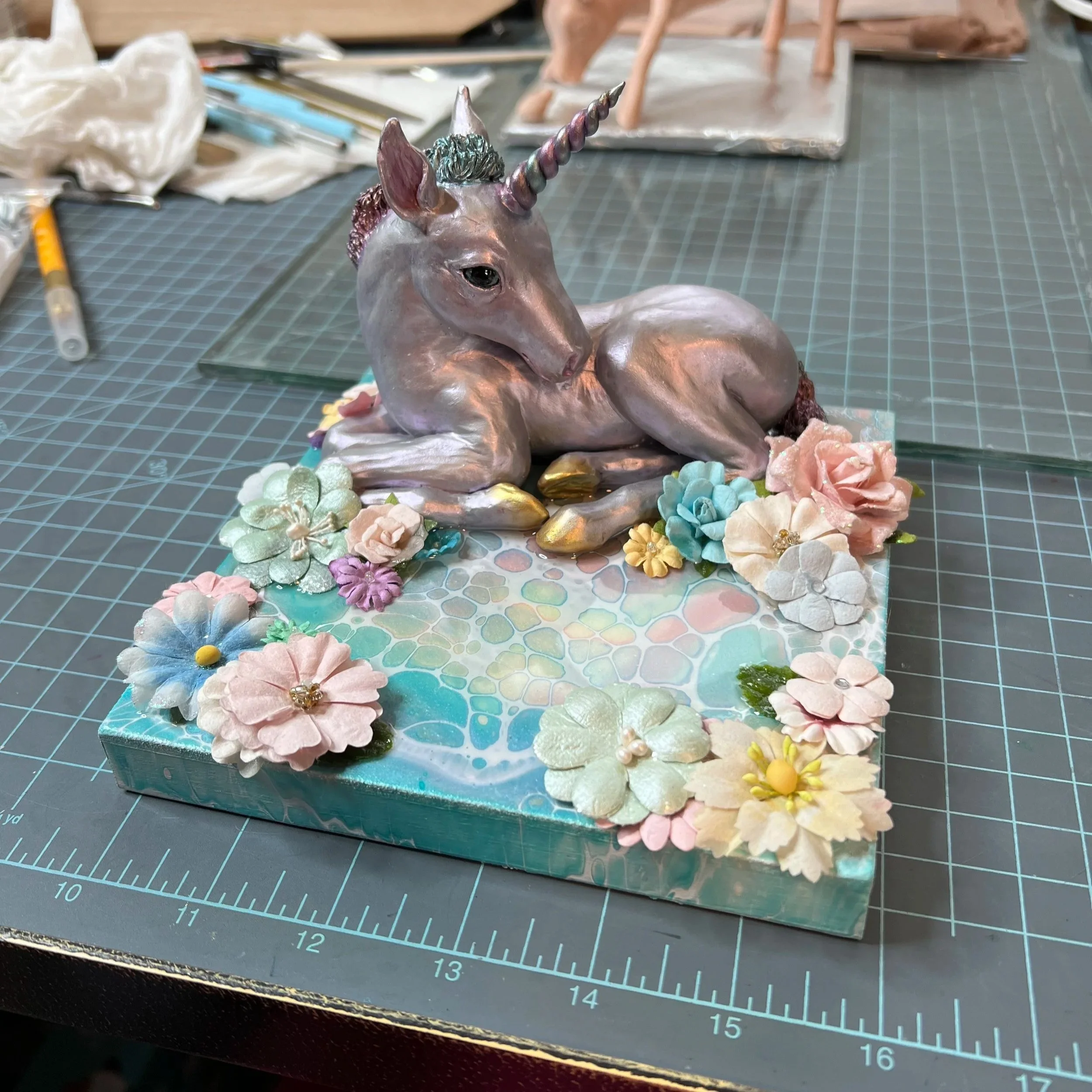 A decorative sculpture of a unicorn lying down on a base decorated with pastel-colored flowers, set on a craft table.