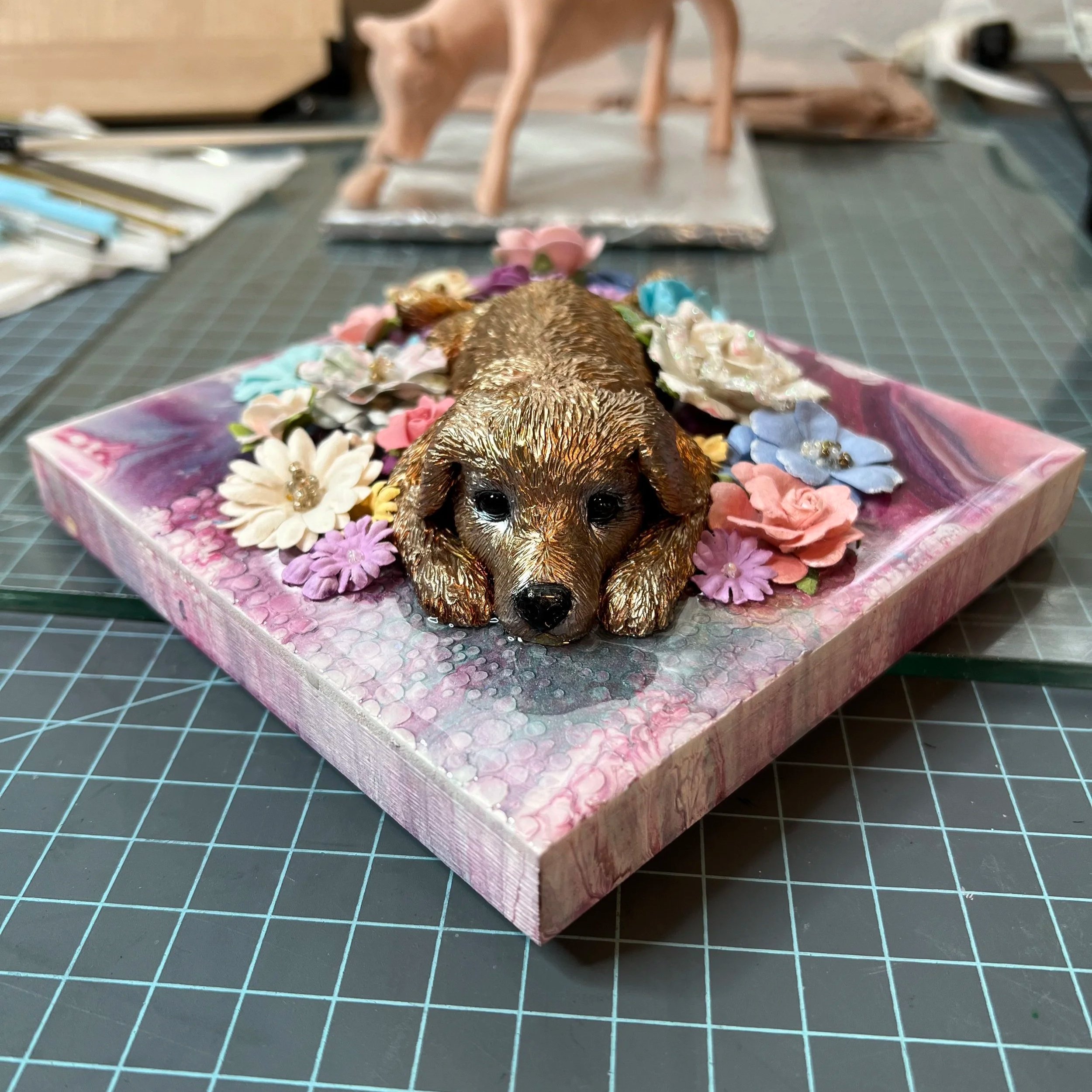 A decorative artwork of a golden retriever puppy lying down among colorful fabric flowers on a textured pink background.