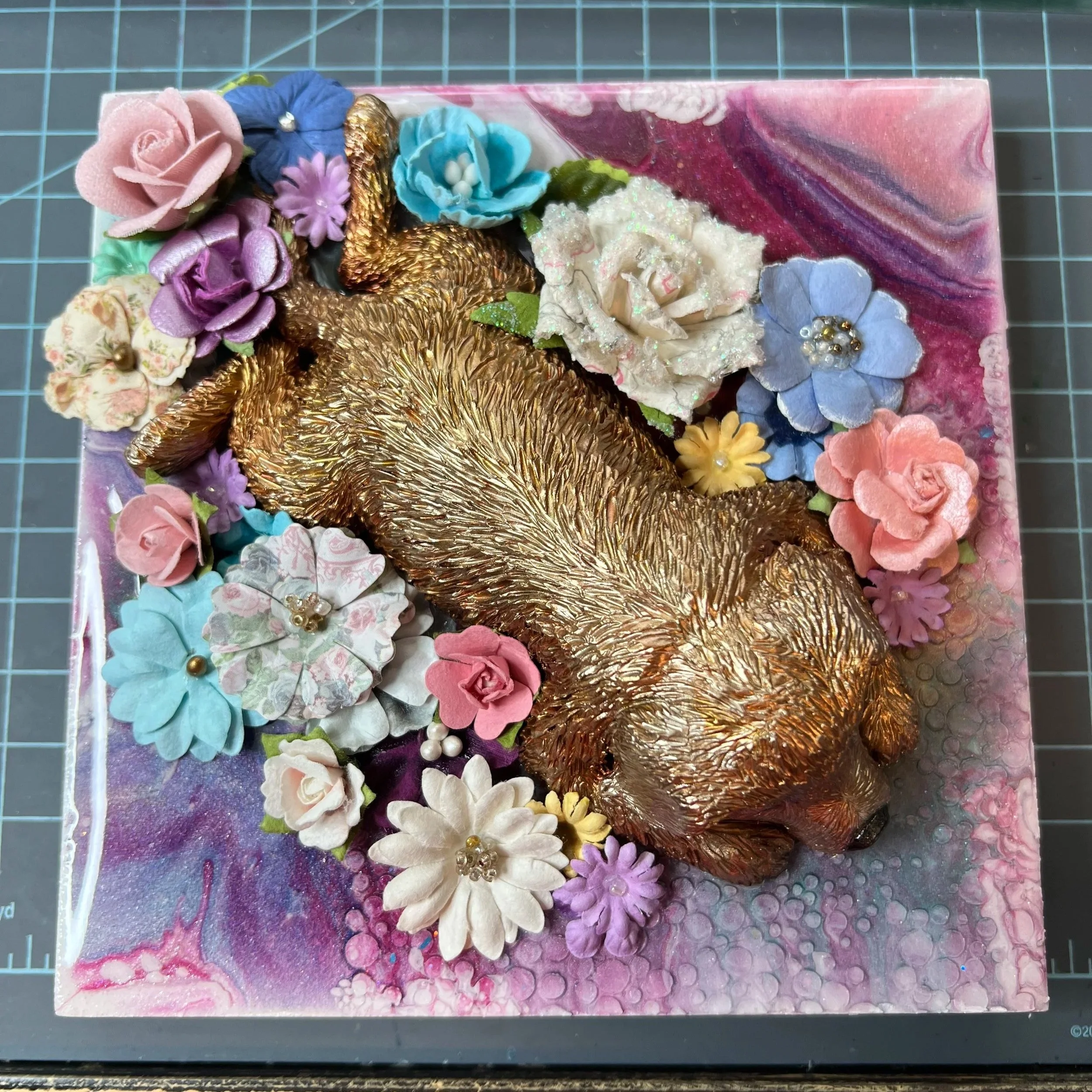 A decorative sculpture of a golden retriever puppy  surrounded by faux flowers on a marbled pink, purple, and white background.