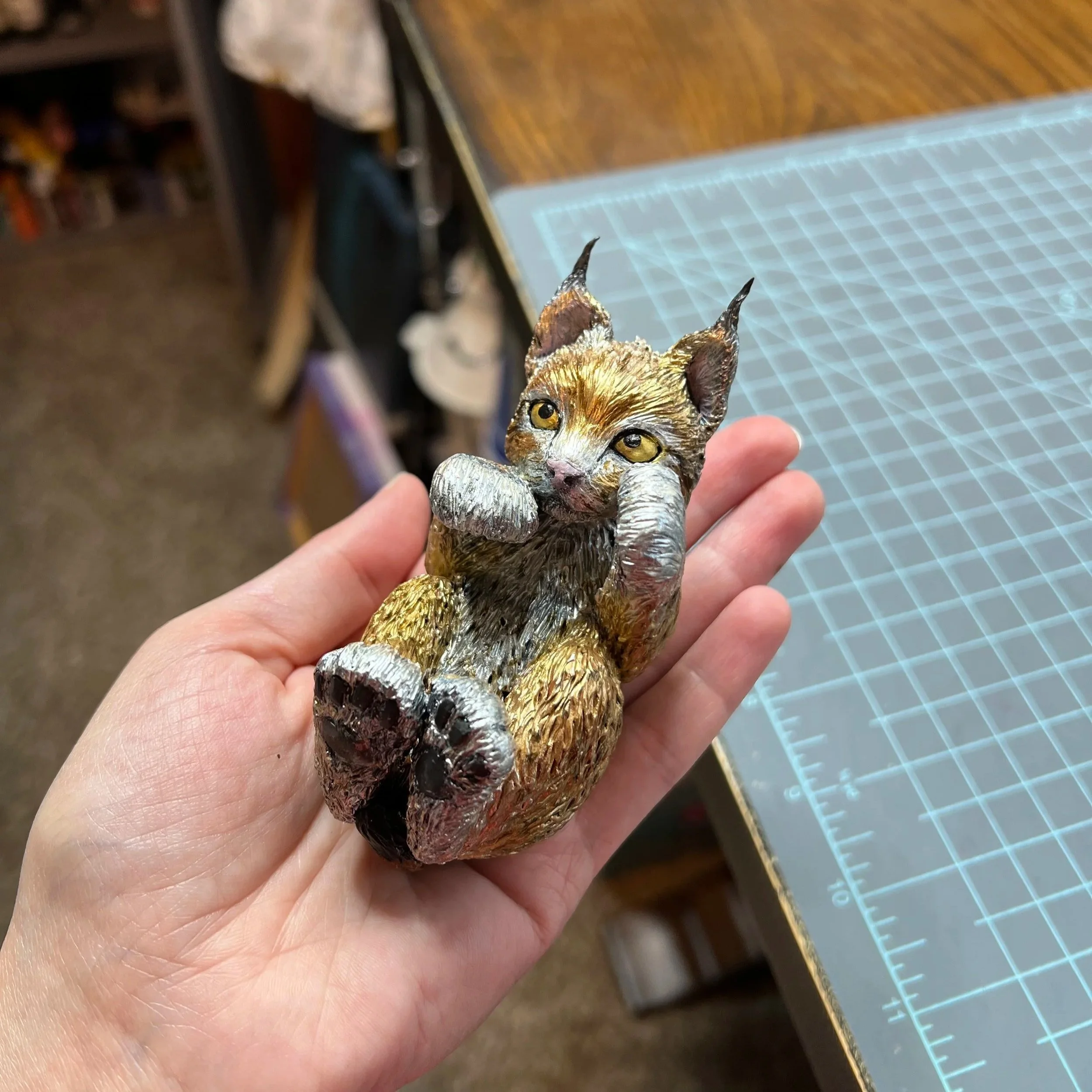 A small, detailed sculpture of a lynx kitten on its back in a playful pose, tail curled up, painted with realistic fur textures and colors.