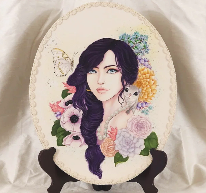 A decorative oval plaque featuring a painted portrait of a woman with long, wavy dark purple hair, surrounded by colorful flowers, a butterfly, and a small white mouse, with a lace-like border.