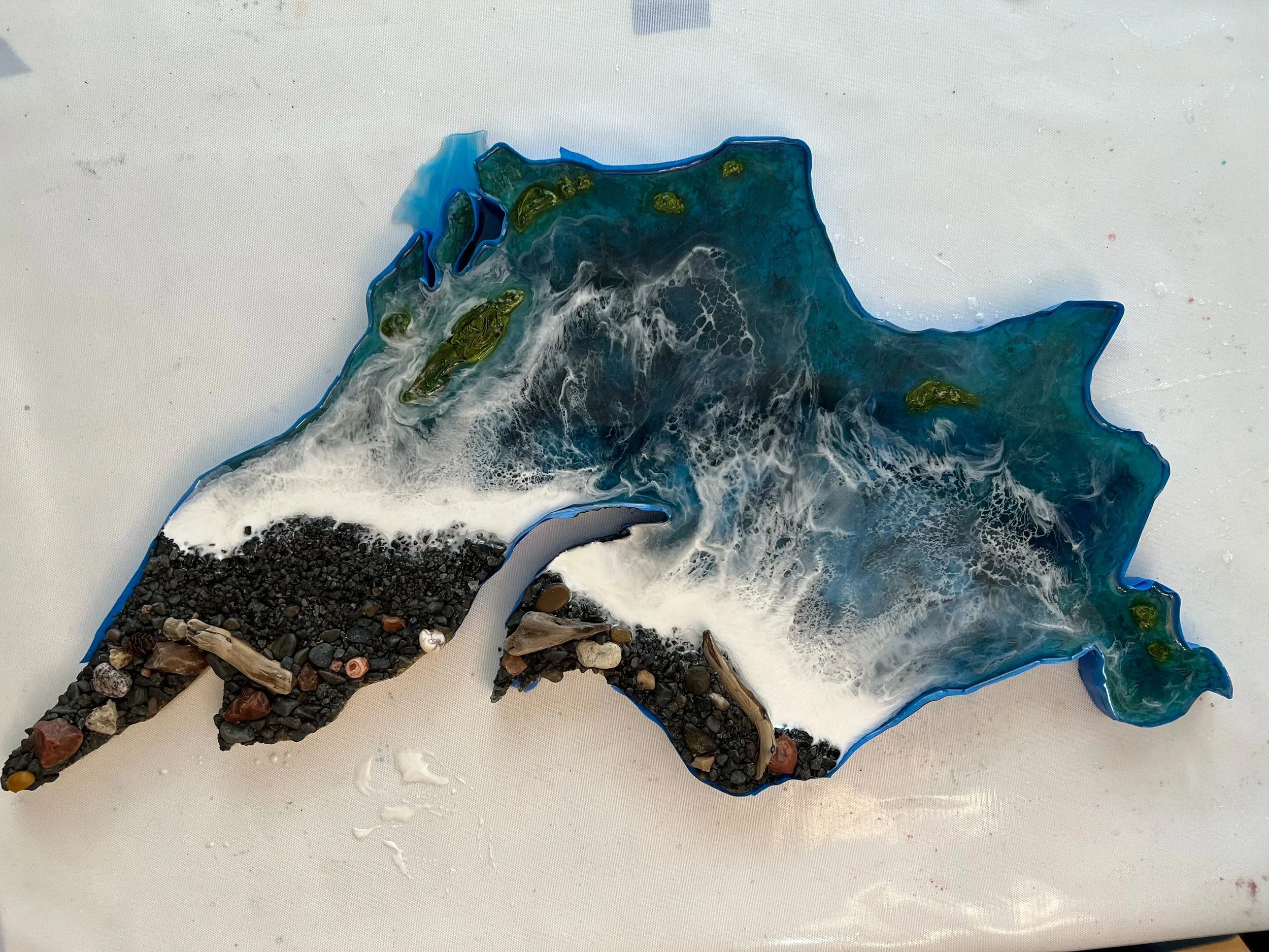 A resin art piece in the shape of the state of Texas, featuring blue, white, and black colors with textured parts resembling mountains and water, and decorative stones and driftwood integrated into the design.