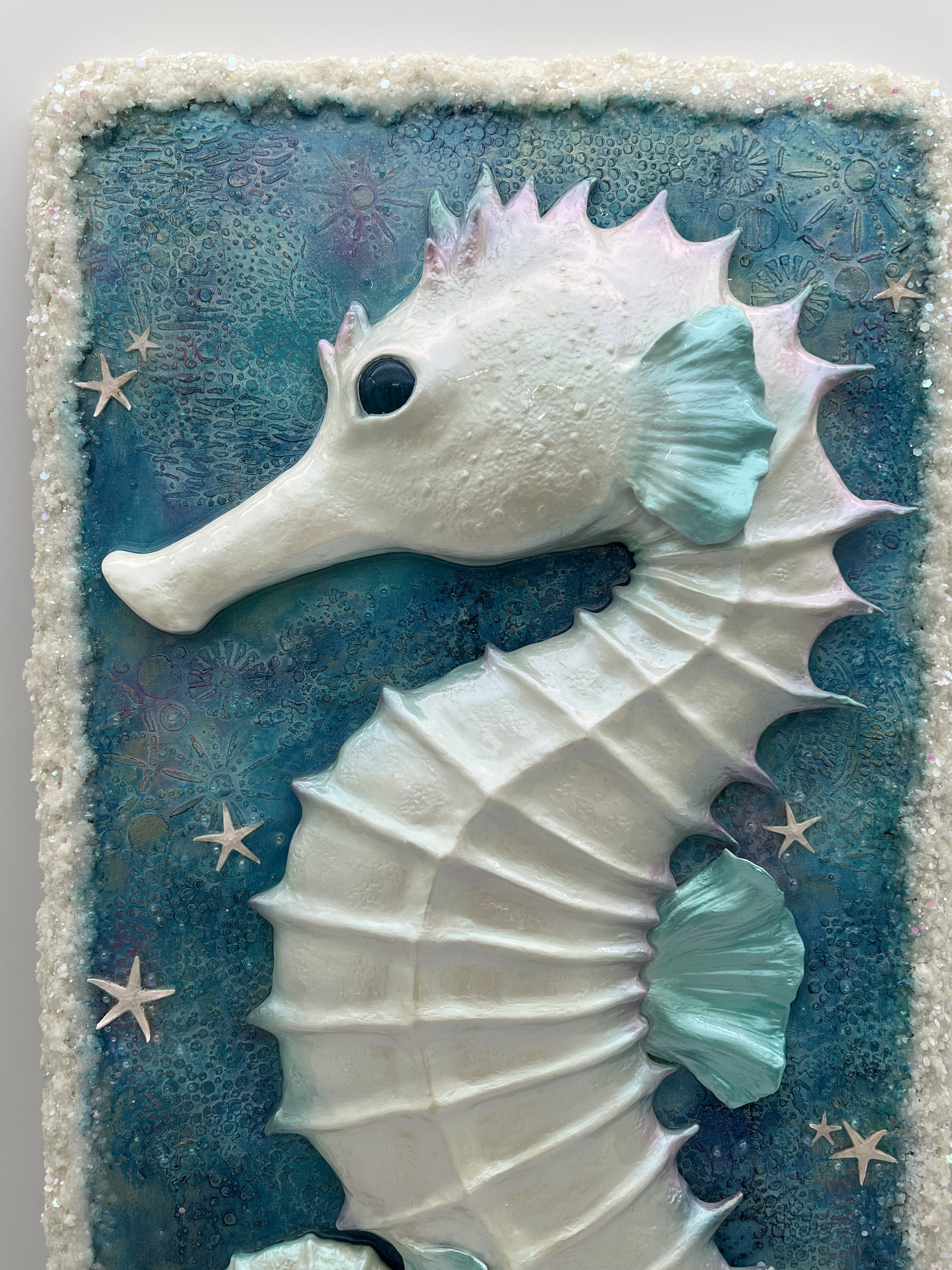 Decorative cake with a seahorse design, featuring a white seahorse with light blue accents on its fins and a textured body, set against a blue background with sea star and shell patterns.