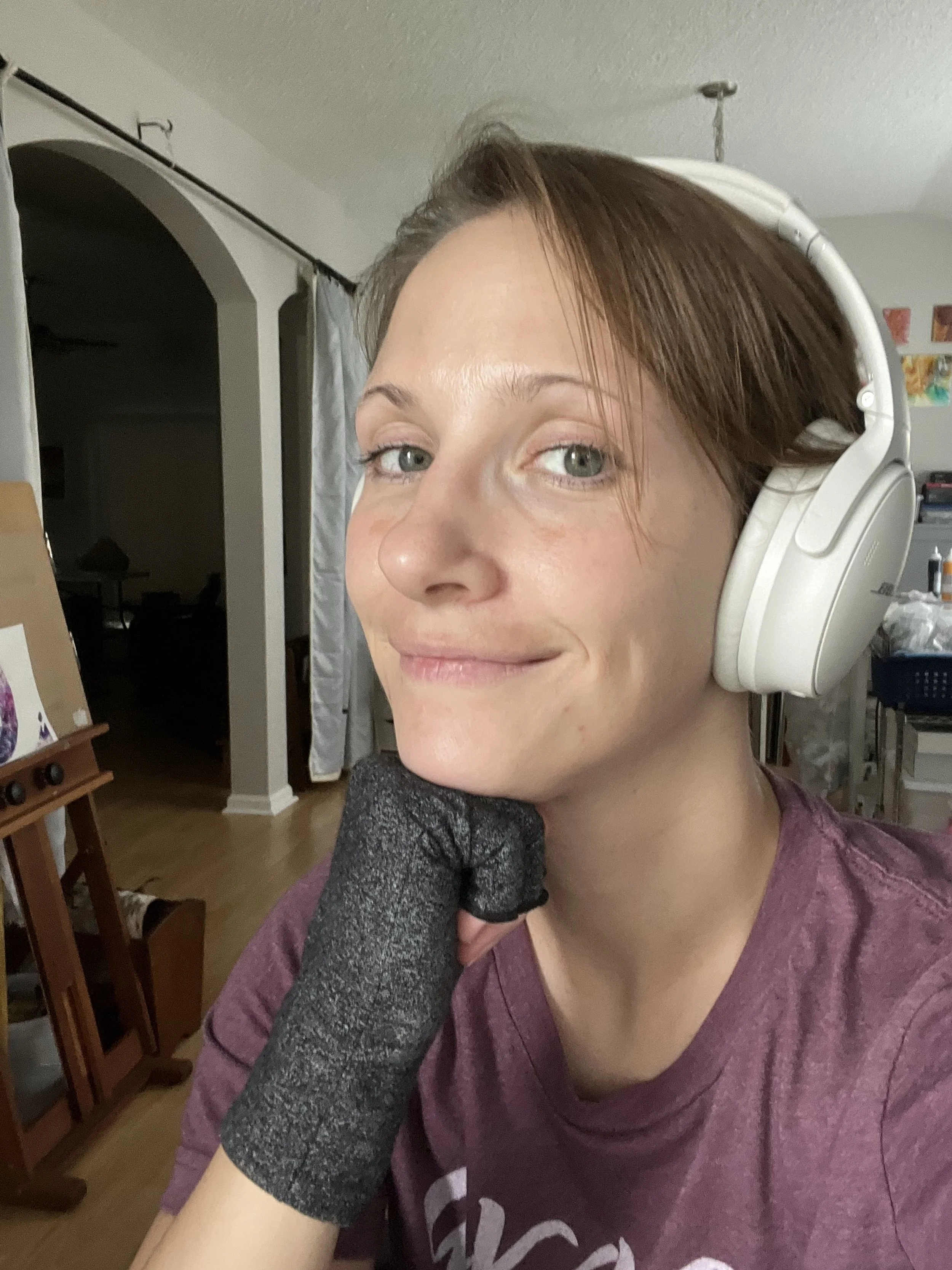 A woman with short brown hair wearing large white headphones, a dark gray glove on her right hand, and a maroon shirt, resting her chin on her hand in a room with a wooden floor and various furniture and art supplies in the background.
