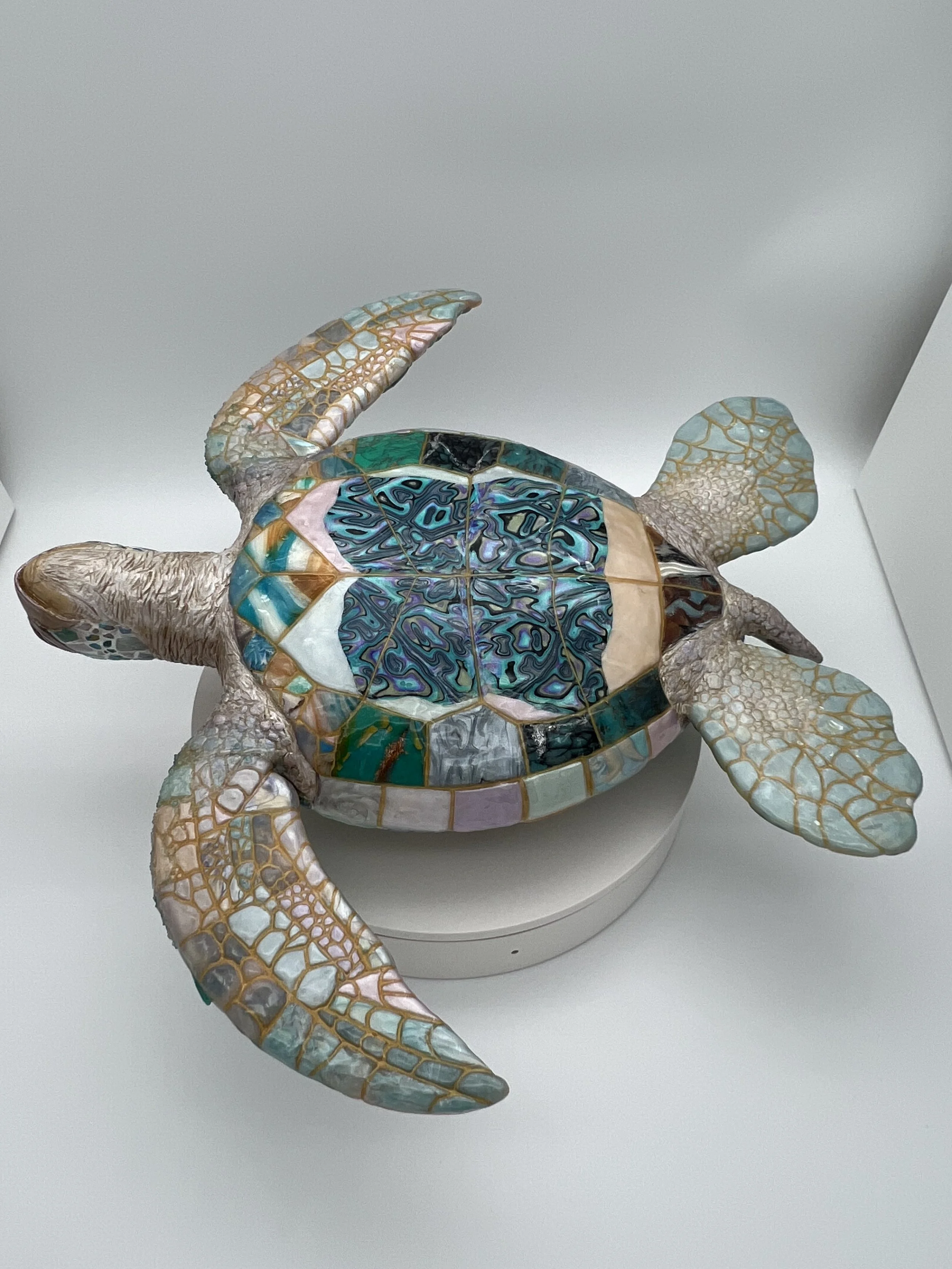 Colorful mosaic turtle sculpture with intricate patterns on its shell and flippers, placed on a white pedestal.