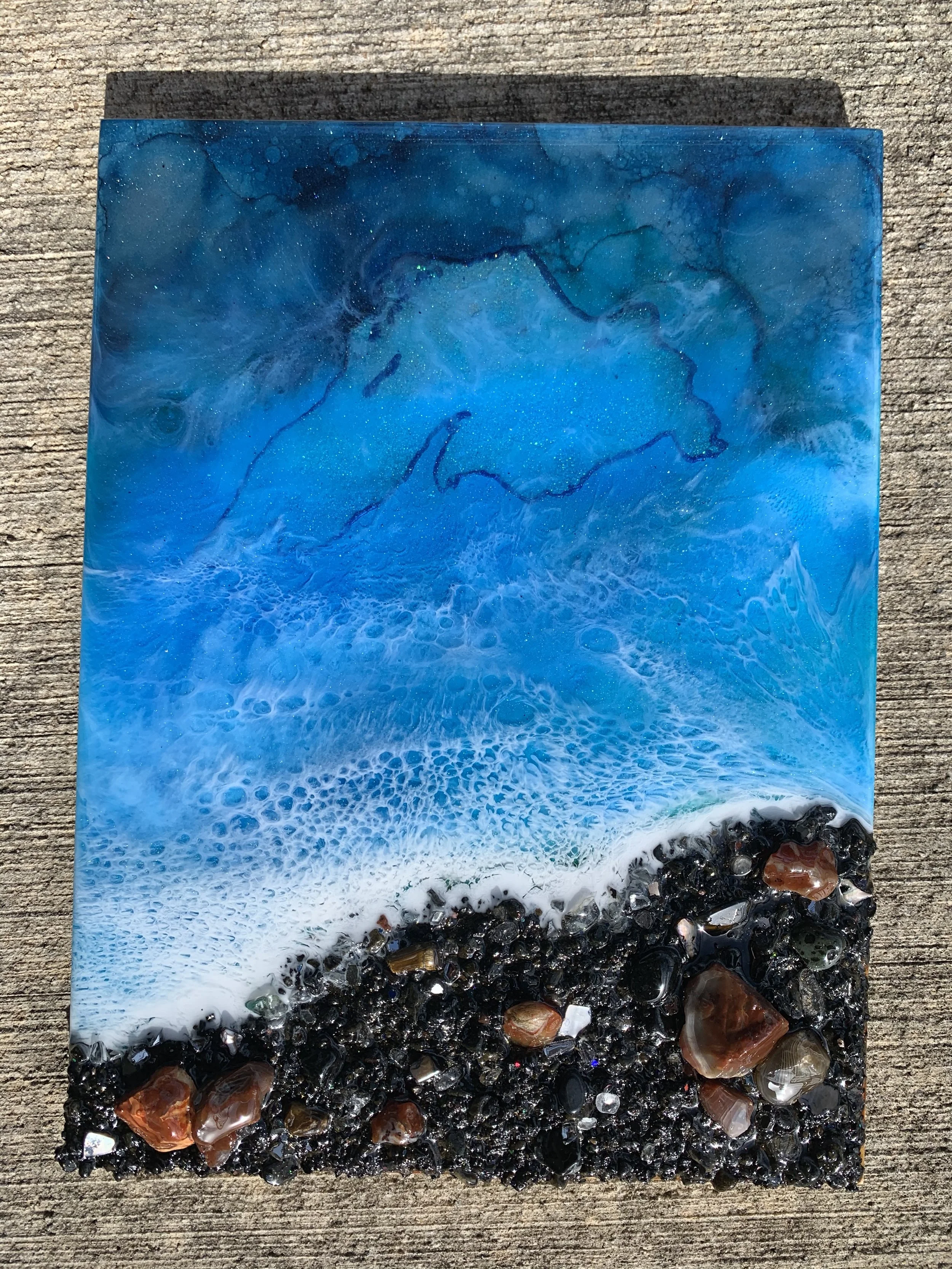 Abstract resin artwork resembling a beach scene with blue ocean, waves, and shoreline with pebbles.