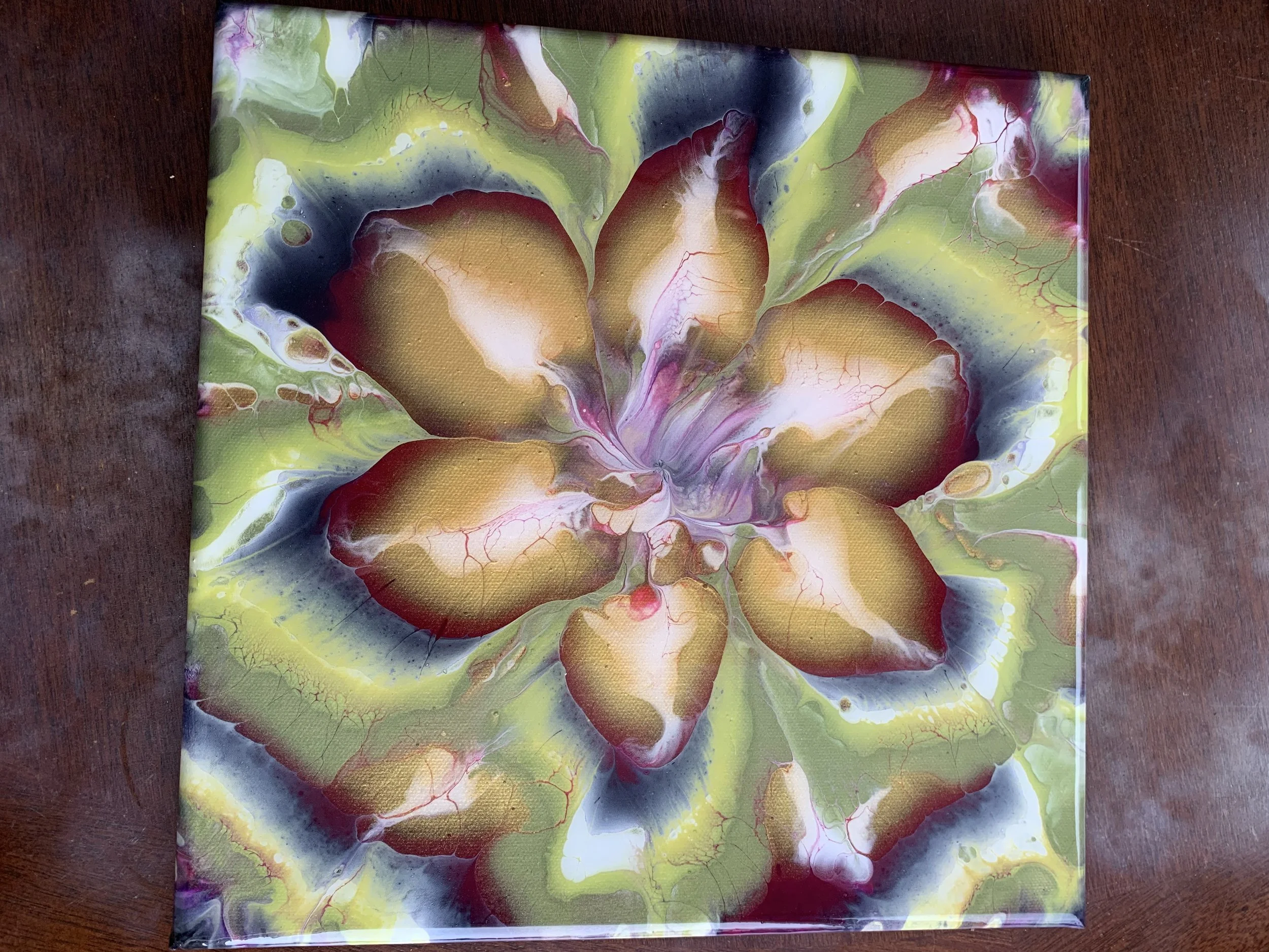 Abstract floral artwork with large, multicolored petals in shades of gold, pink, purple, black, and green, with a swirling pattern and detailed veining, on a wooden surface.