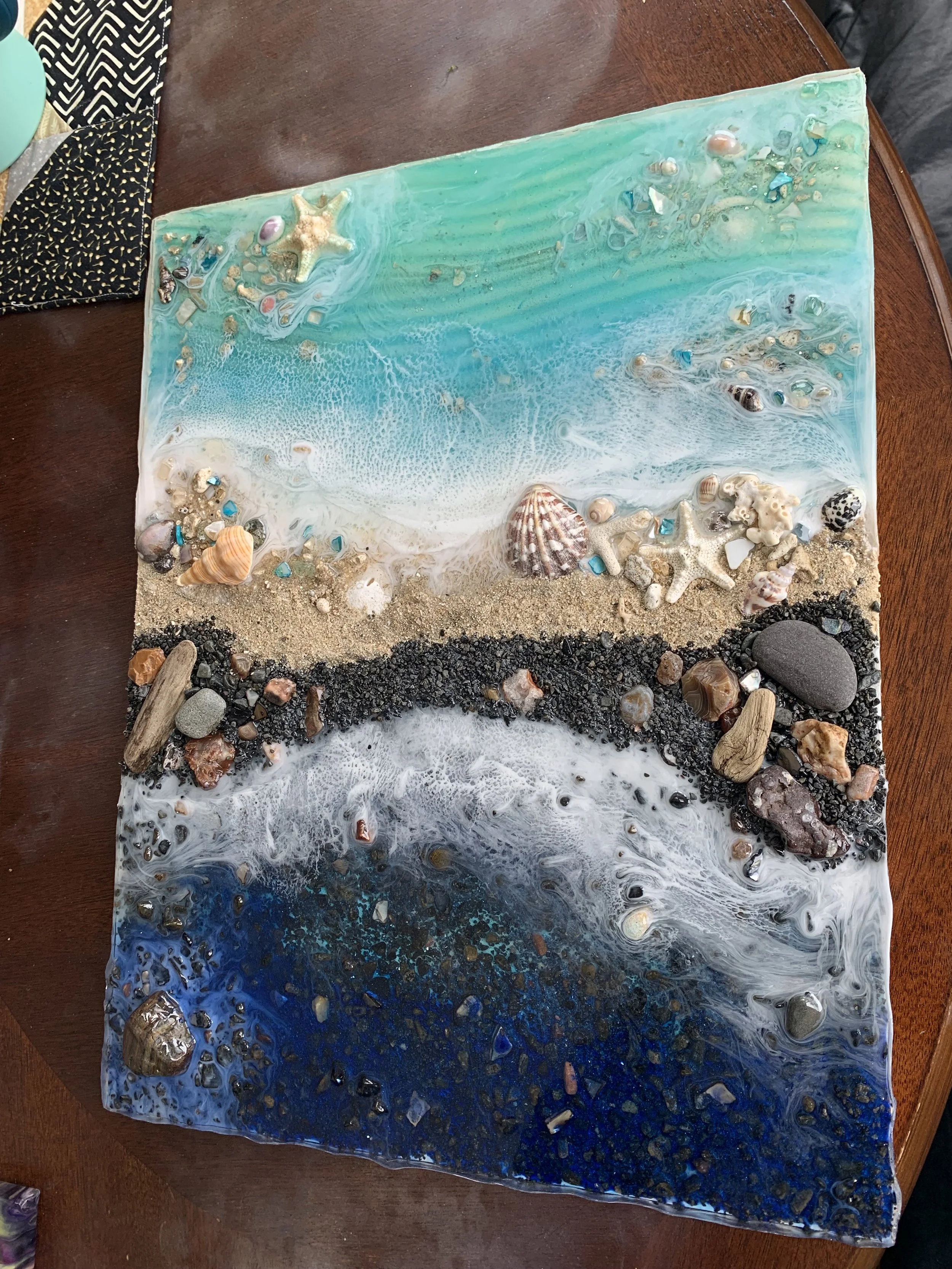 Mixed media artwork depicting a beach scene divided into three sections: ocean at the top with shades of blue and white foam, a middle sandy shoreline with seashells and small decorative stones, and a lower section with dark blue and black colors rep