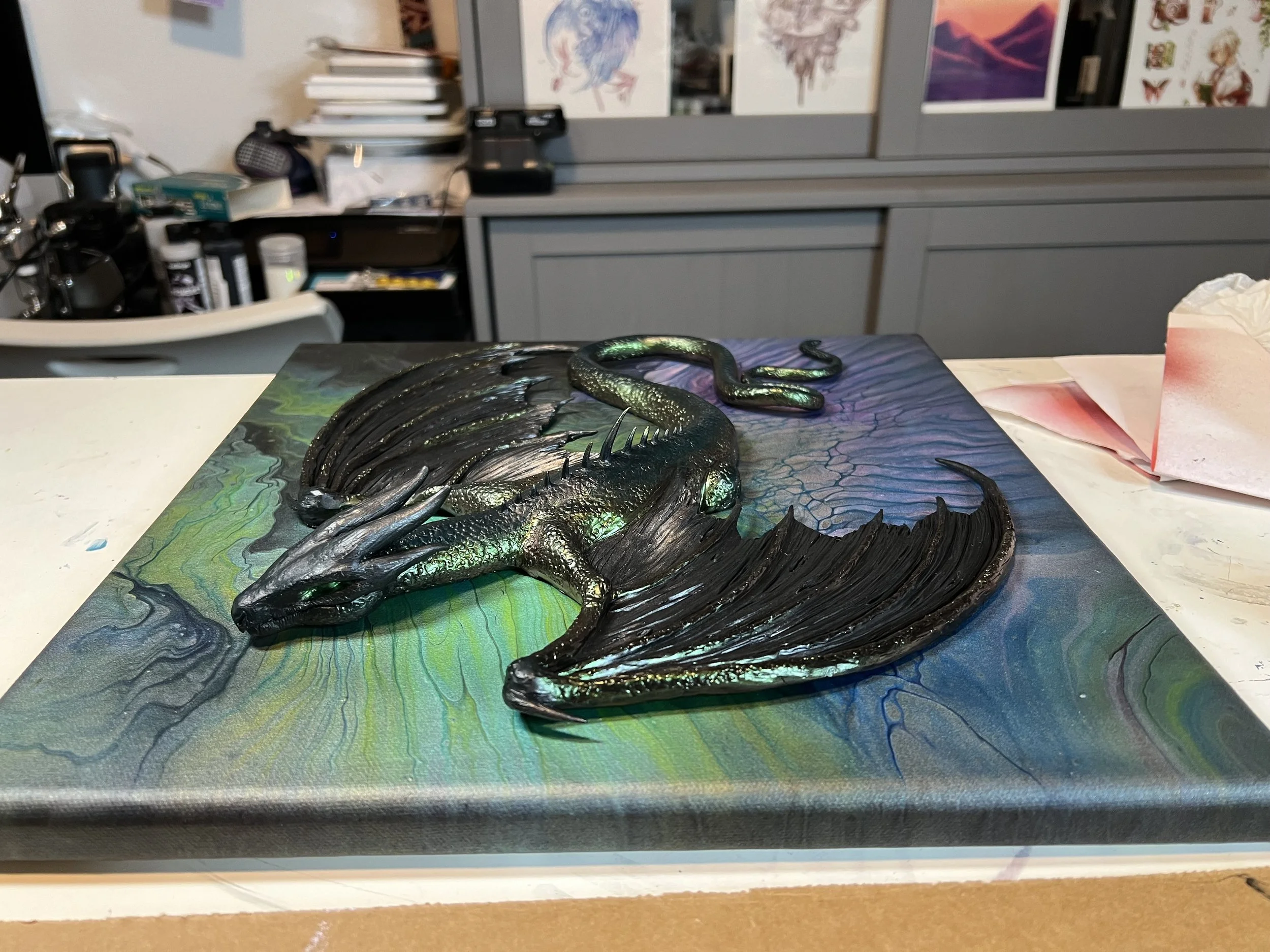 A 3D mixed-media artwork of a dragon laying on a textured, colorful background. The dragon has dark scales with iridescent green accents, and its wings are large and textured.