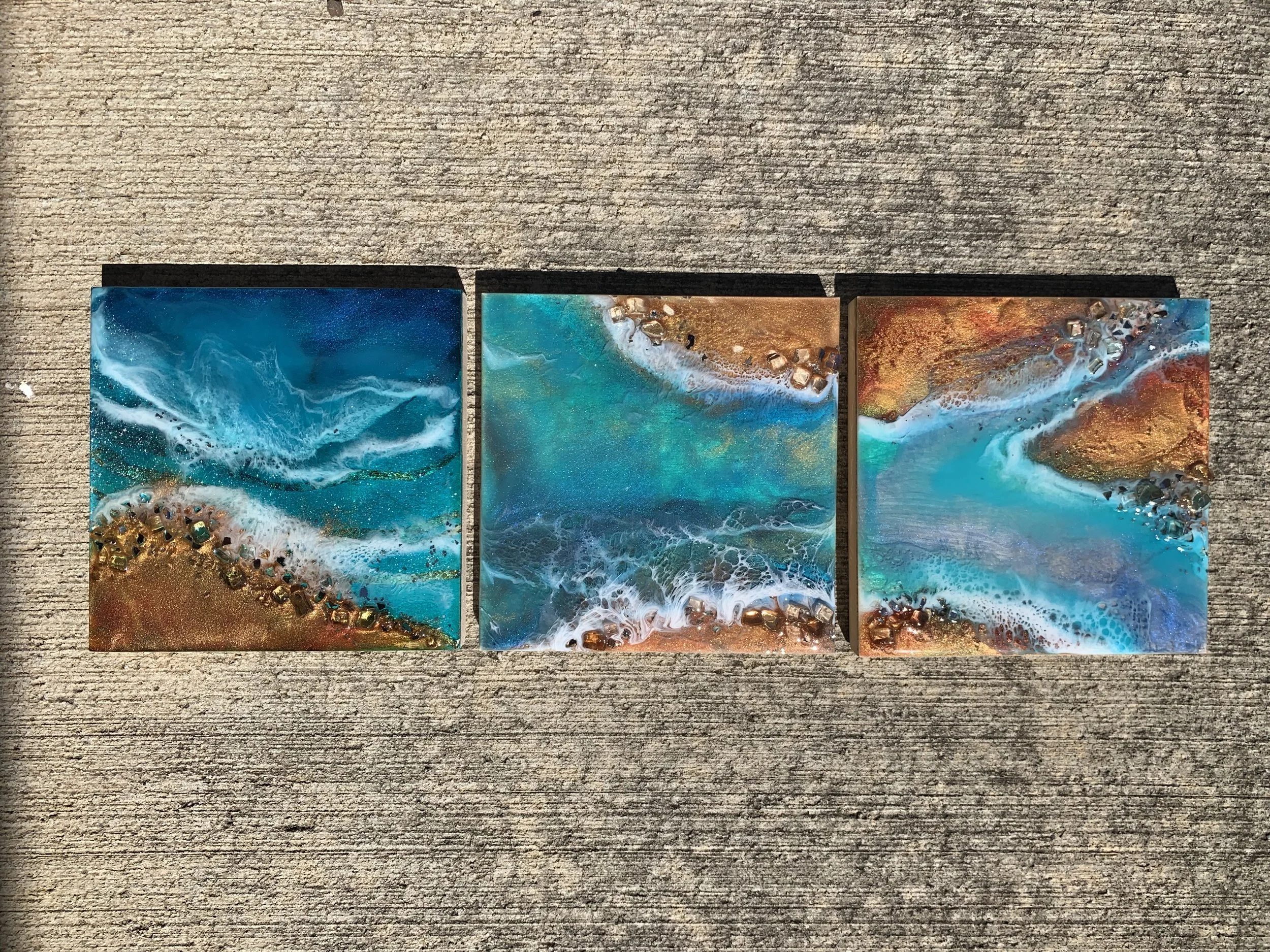 Three rectangular abstract paintings resembling ocean waves with blue, white, gold, and copper colors, placed on a textured gray surface.