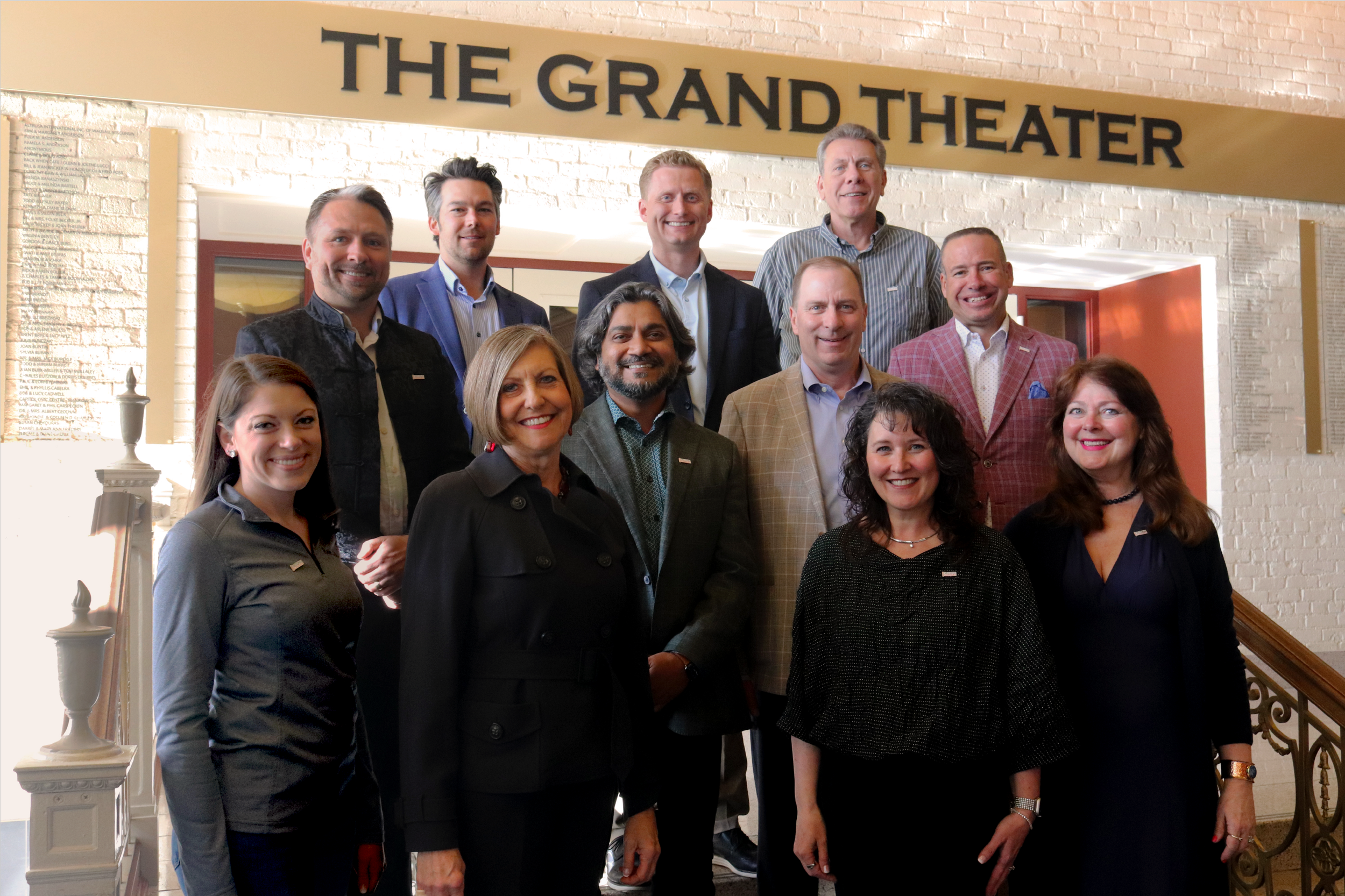 A group of thirteen professionals posing in front of a sign that reads 'The Grand Theater' inside a building with brick walls and a staircase.