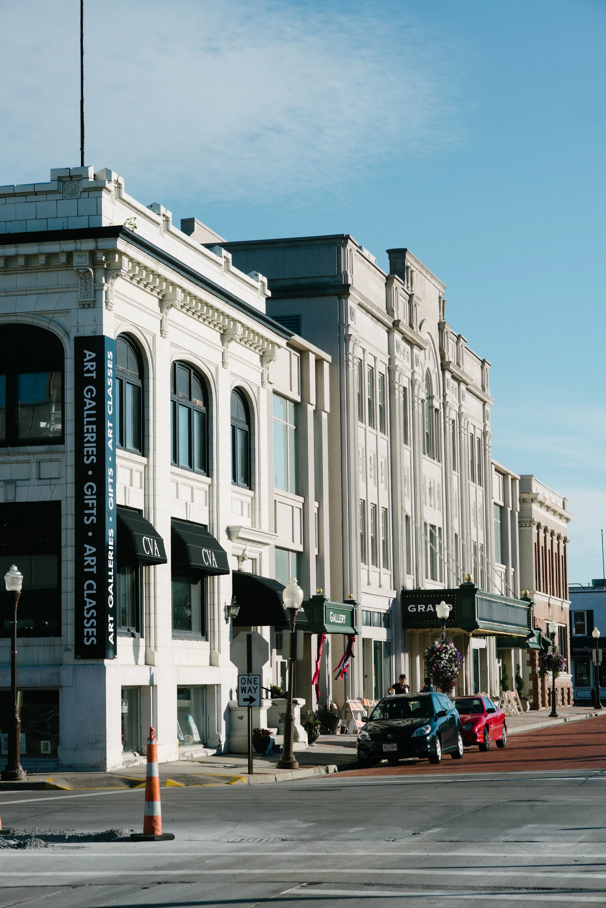 Street view of historic buildings with shops, art galleries, and cars parked along the curb, under a blue sky.