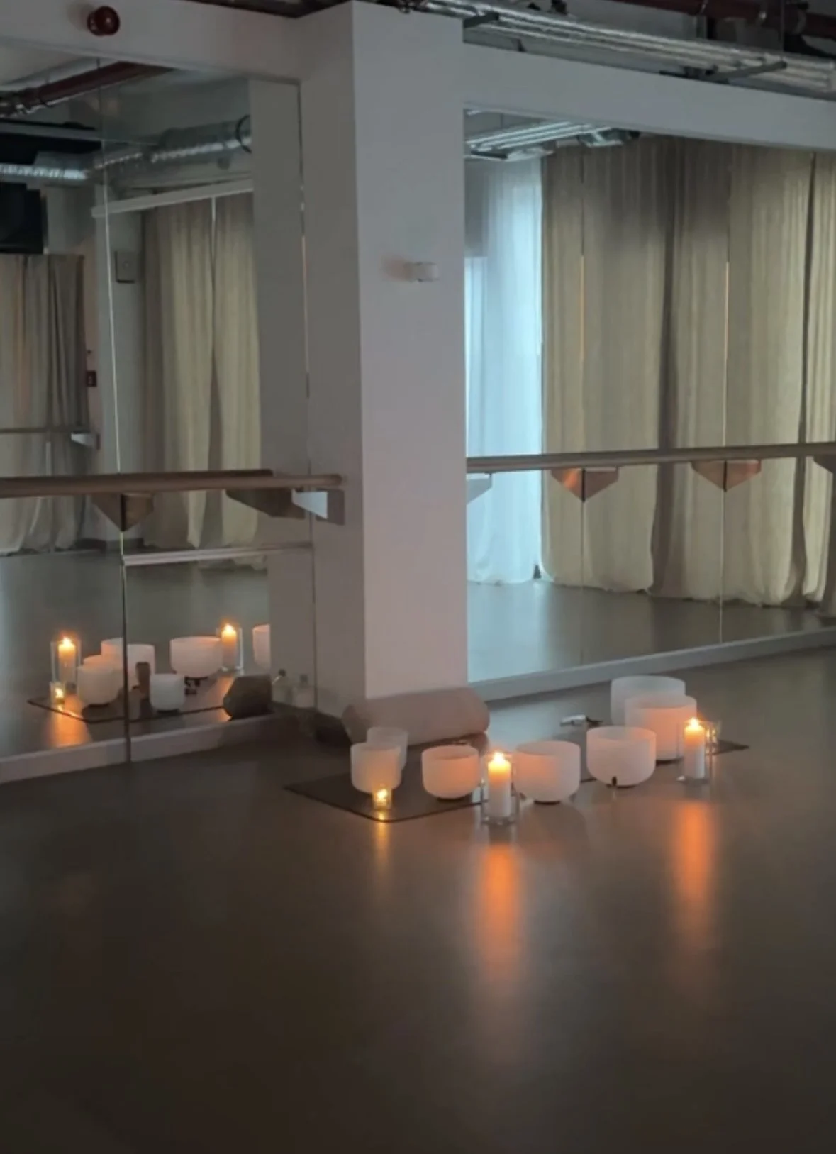 A dance studio with a wooden floor, large wall mirrors, and beige curtains. Candles are arranged on the floor near the wall, creating a warm, ambient glow.