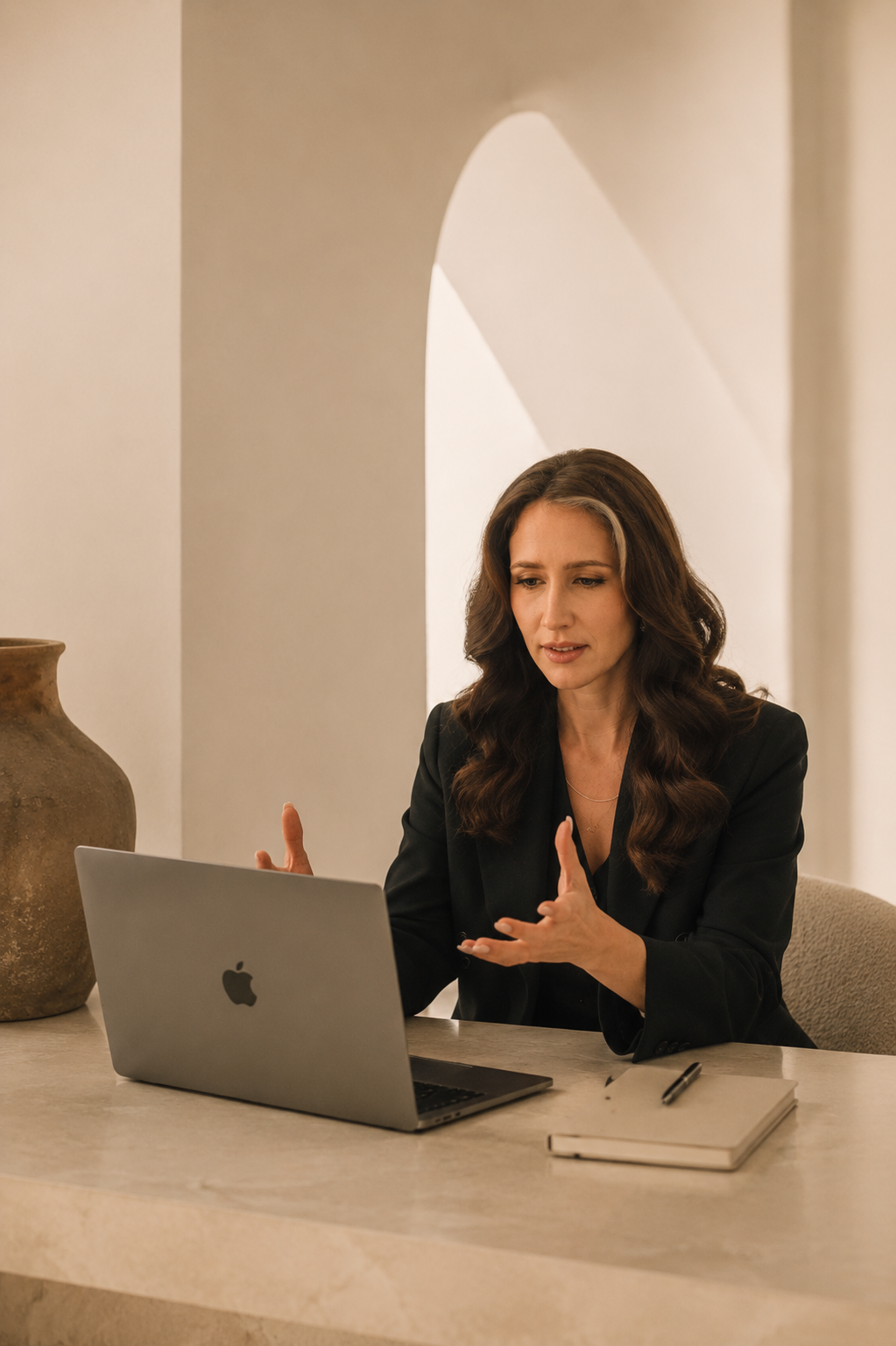 A woman with long wavy brown hair dressed in a black blazer, seated at a beige table with a silver MacBook laptop, notebook, and pen, gesturing with her hands during a discussion or presentation in a neutral-toned room with a large decorative vase on the side.