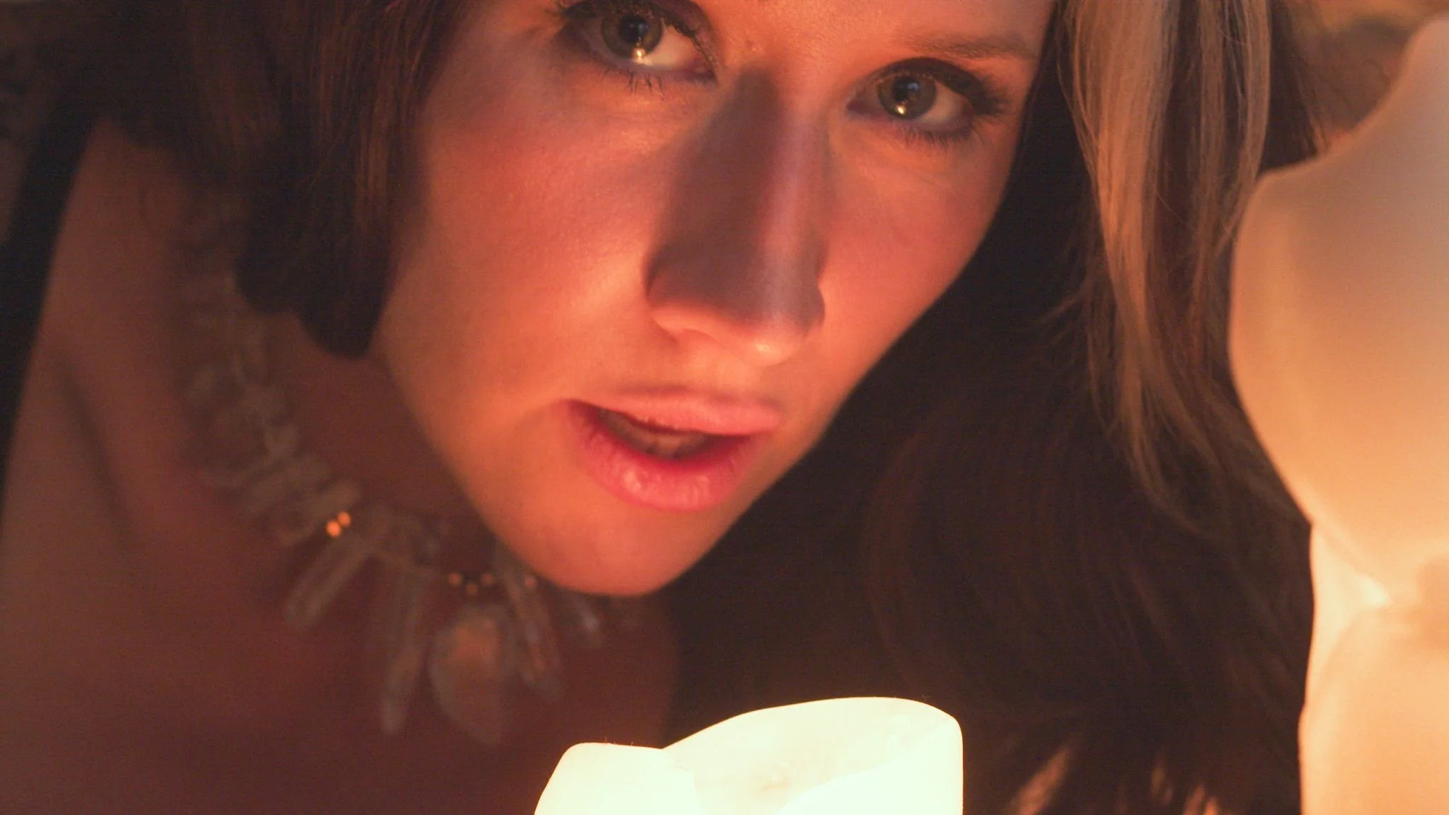 A woman with long brown hair looking at a lit candle, with a curious or contemplative expression on her face.