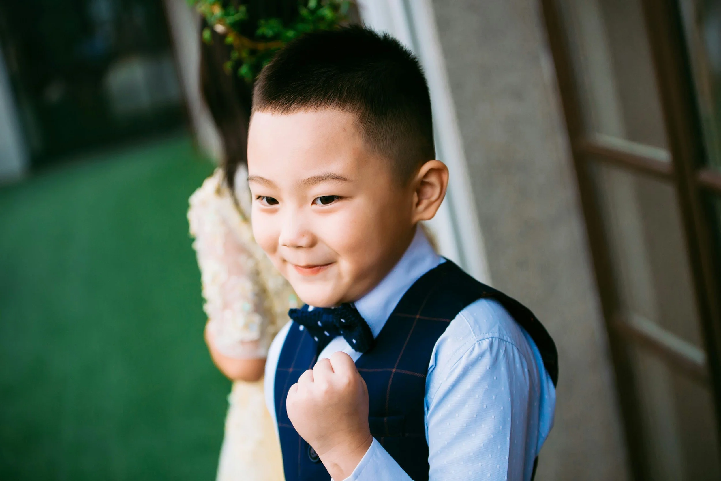 Smiling young boy in formal attire with a vest and bow tie, and a handshake gesture, standing outdoors near a building.