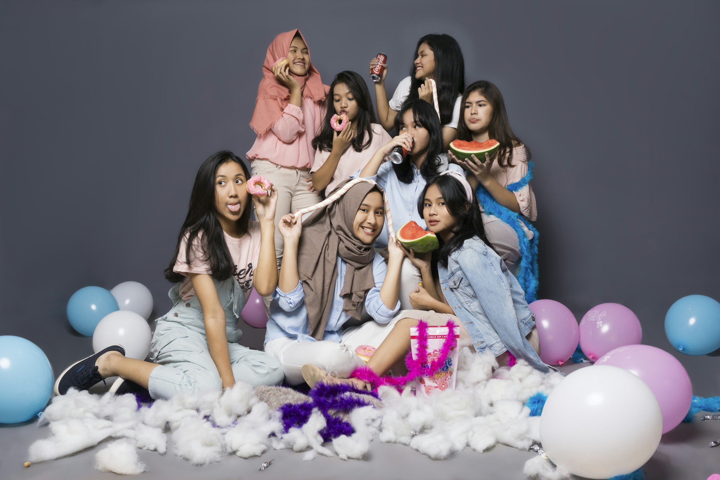 Group of nine young girls celebrating a party with balloons, cake, and cotton decoration on a gray background.
