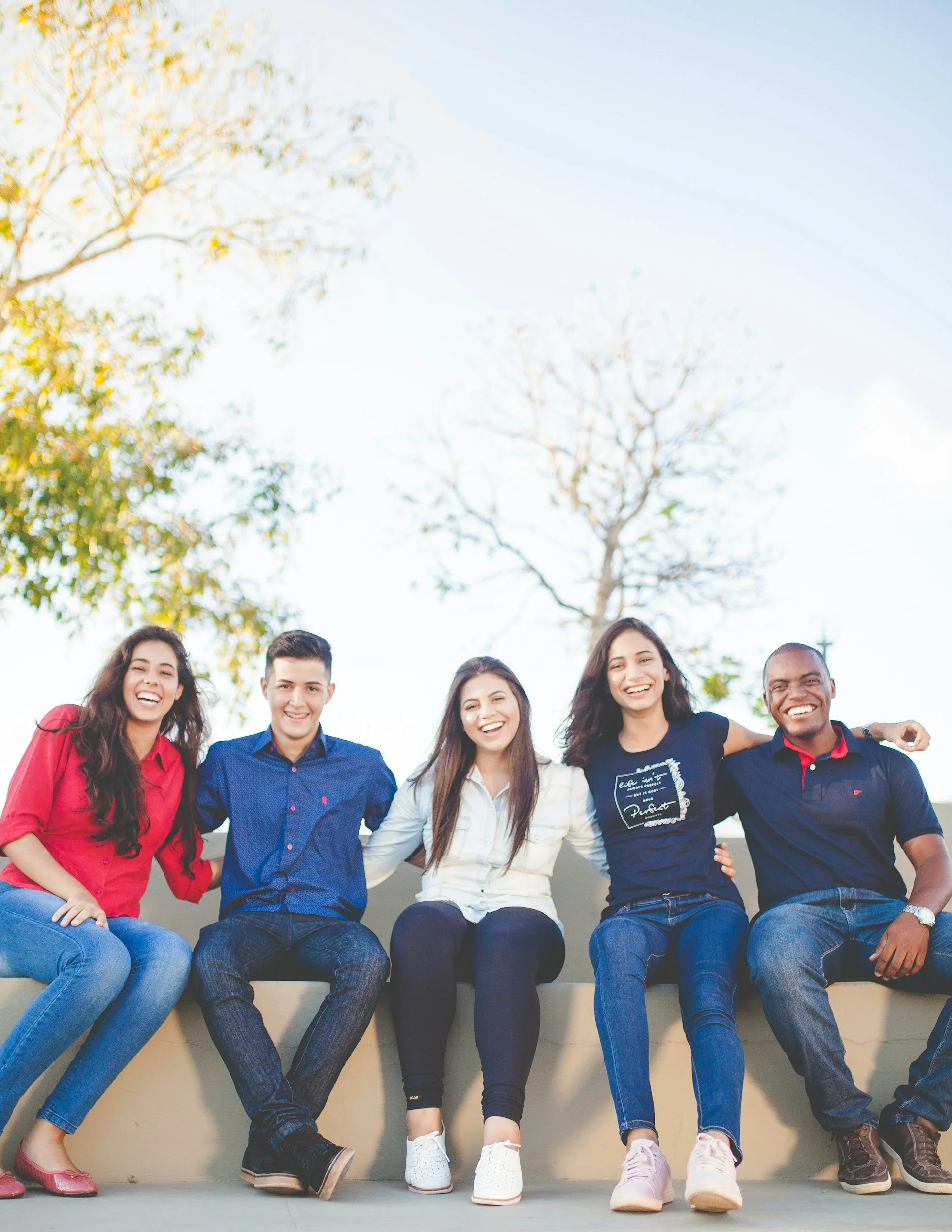 Group of five young adults sitting together outside, smiling, with trees and blue sky in the background.