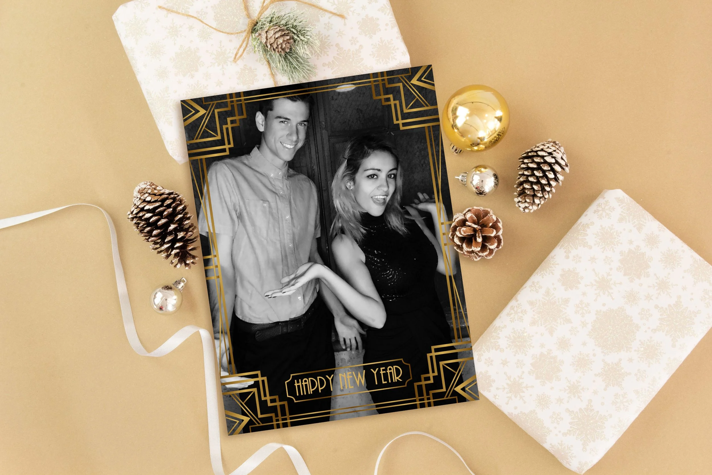 A black and white photo of a young man and woman celebrating New Year's, surrounded by holiday decorations, including pinecones and shiny ornaments, on a beige background with wrapped gifts.