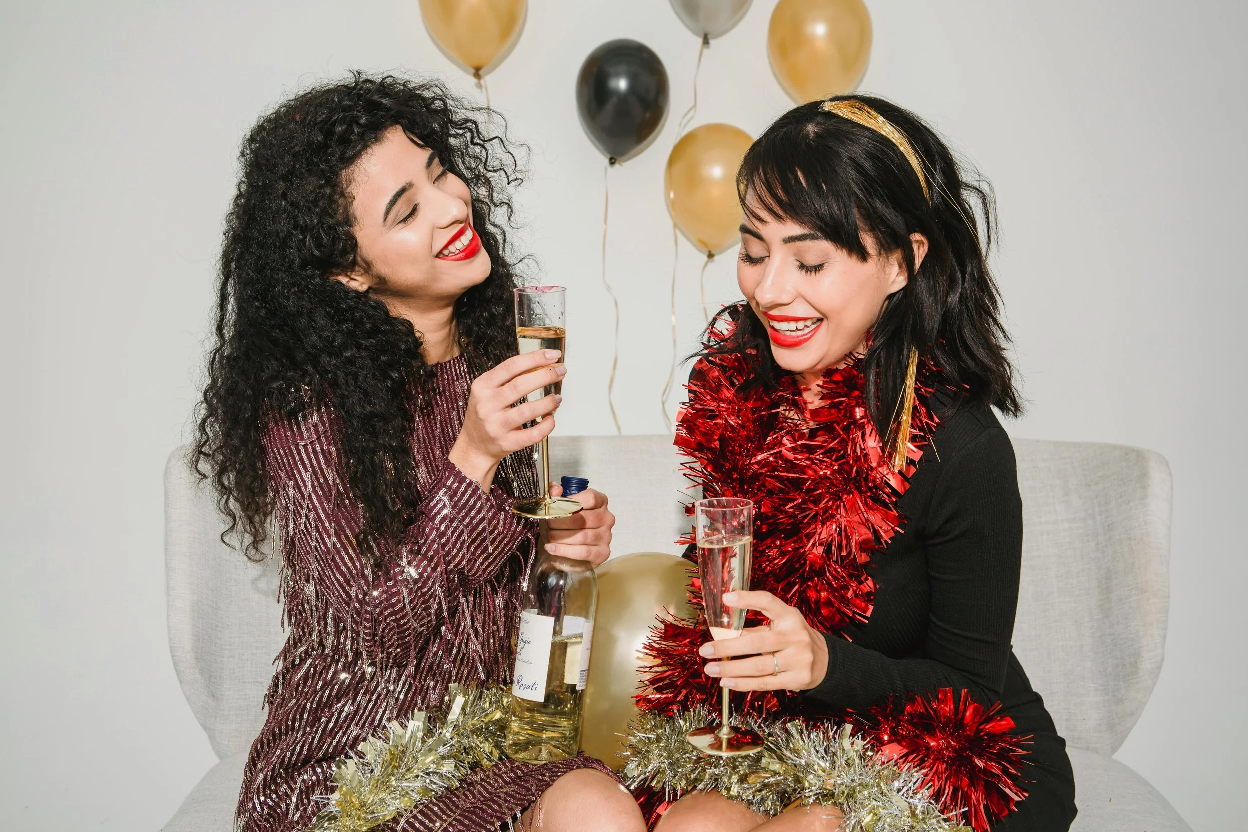 Two women celebrating with drinks, wearing festive decorations, with balloons in the background.