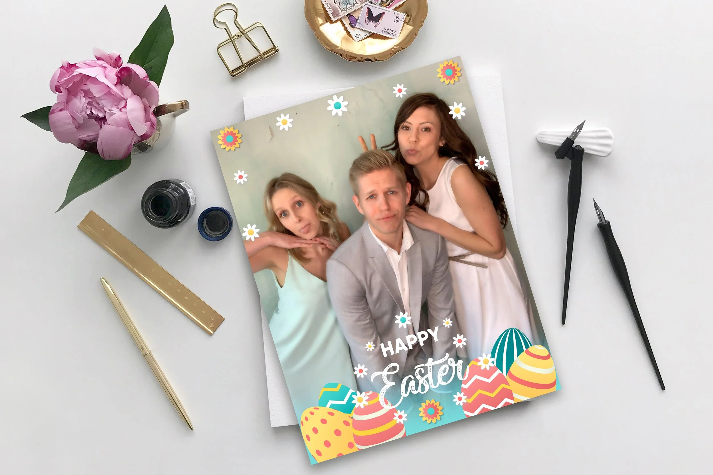 A printed photograph with three people making funny faces. The photo has floral and Easter egg decorations with a "Happy Easter" message. The photograph is on a white surface, surrounded by a pink flower in a mug, gold and black pens, ink bottles, a gold ruler, a pen knife, a bowl of postage stamps, and a letter opener.